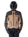 Alpinestars AMT-8 Stretch Drystar XF jas, adventure touring motorjas, donkerkhaki en donkerbruin, voorzien van oranje Astars-logo en zwarte ritsen, gedragen met zwarte adventure-helm en handschoenen, volledige motoruitrusting