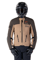 Alpinestars AMT-8 Stretch Drystar XF jas, adventure touring motorjas, donkerkhaki en donkerbruin, voorzien van oranje Astars-logo en zwarte ritsen, gedragen met zwarte adventure-helm en handschoenen, volledige motoruitrusting