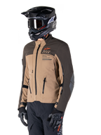 Alpinestars AMT-8 Stretch Drystar® XF Jas, adventure motorjas, donkerbruin en donker khaki, gedragen met een zwarte off-road helm en crossbril, voorzien van Alpinestars Motion Technology-branding en ventilatieopeningen met ritssluiting.
