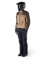 Adventure-motorrijder in volledige uitrusting, Alpinestars AMT-8 Stretch Drystar® XF Jas in donkerbruin en donkerkhaki, gecombineerd met zwarte adventure-broek, zwarte helm, handschoenen en laarzen, voorzien van oranje AMT-branding en technische ventilatieritsen voor touring over lange afstanden