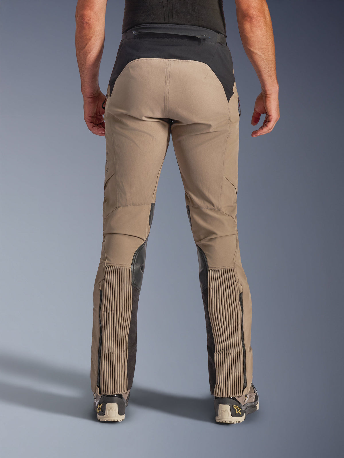 Alpinestars AMT-8 Stretch Drystar® XF broek, Walnut Black, achteraanzicht op model, tan stretch textiel chassis met zwart taille-inzetstuk en kuitpanelen, voorzien van grote accordeon-stretchgedeelten op de onderbenen, beenopeningen met ritssluiting en adventure-touring pasvorm