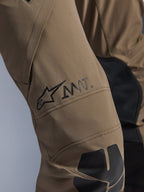 Alpinestars AMT-8 Stretch Drystar® XF-broek in Walnut Black, close-up van de broekspijp met het zwarte AMT-logo en de Alpinestars-ster, hoogwaardig stretchtextiel met zwarte panelen, technische adventure-motoruitrusting