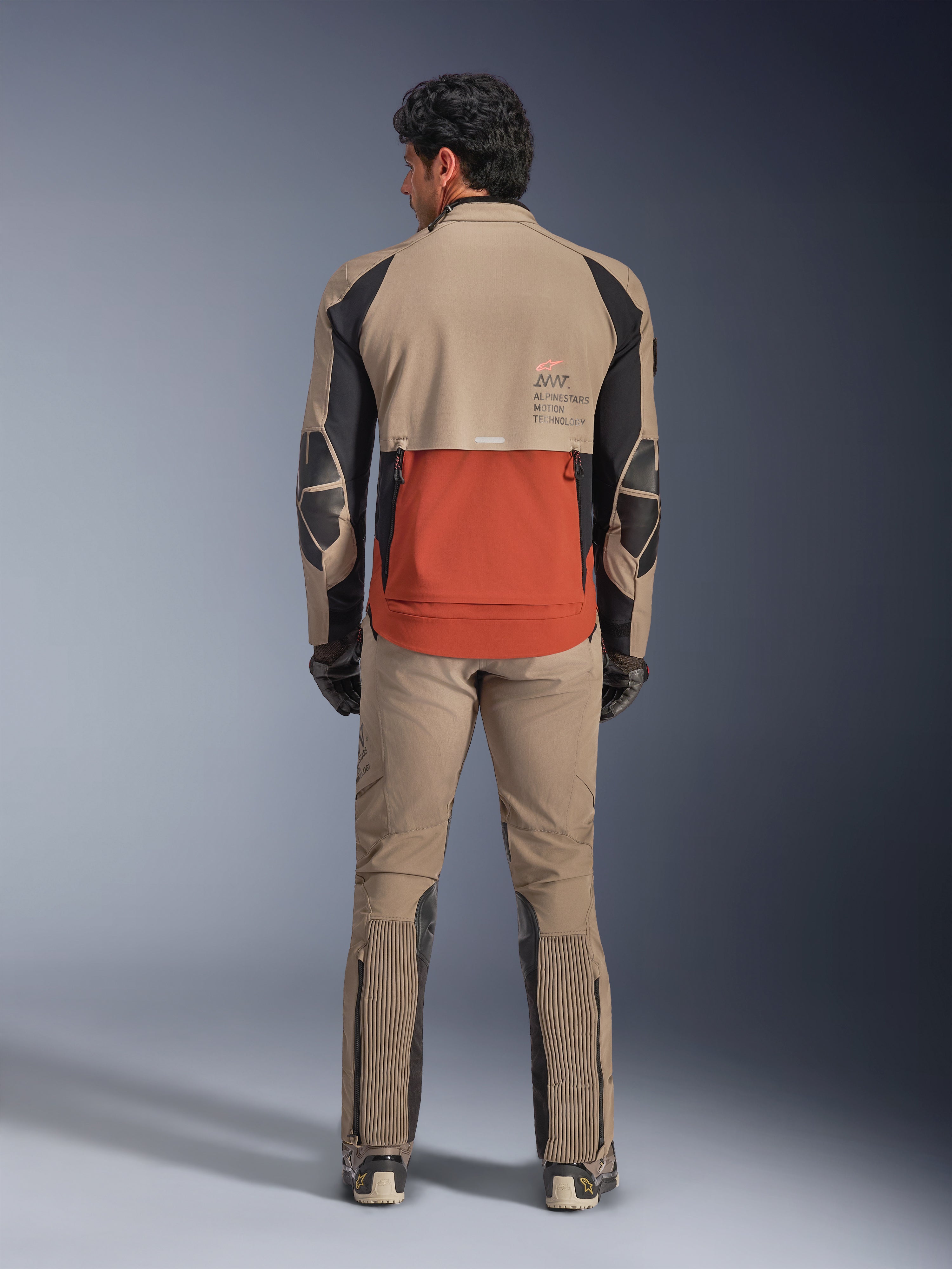 Alpinestars AMT-8 Stretch Drystar® XF Jas, Walnut Rust Black, achteraanzicht op een rijder met bijpassende adventure broek en handschoenen, voorzien van een opvallend roestkleurig achterpaneel, walnootkleurige bovendelen en zwarte stretchaccenten, ontworpen voor lange afstandsritten.