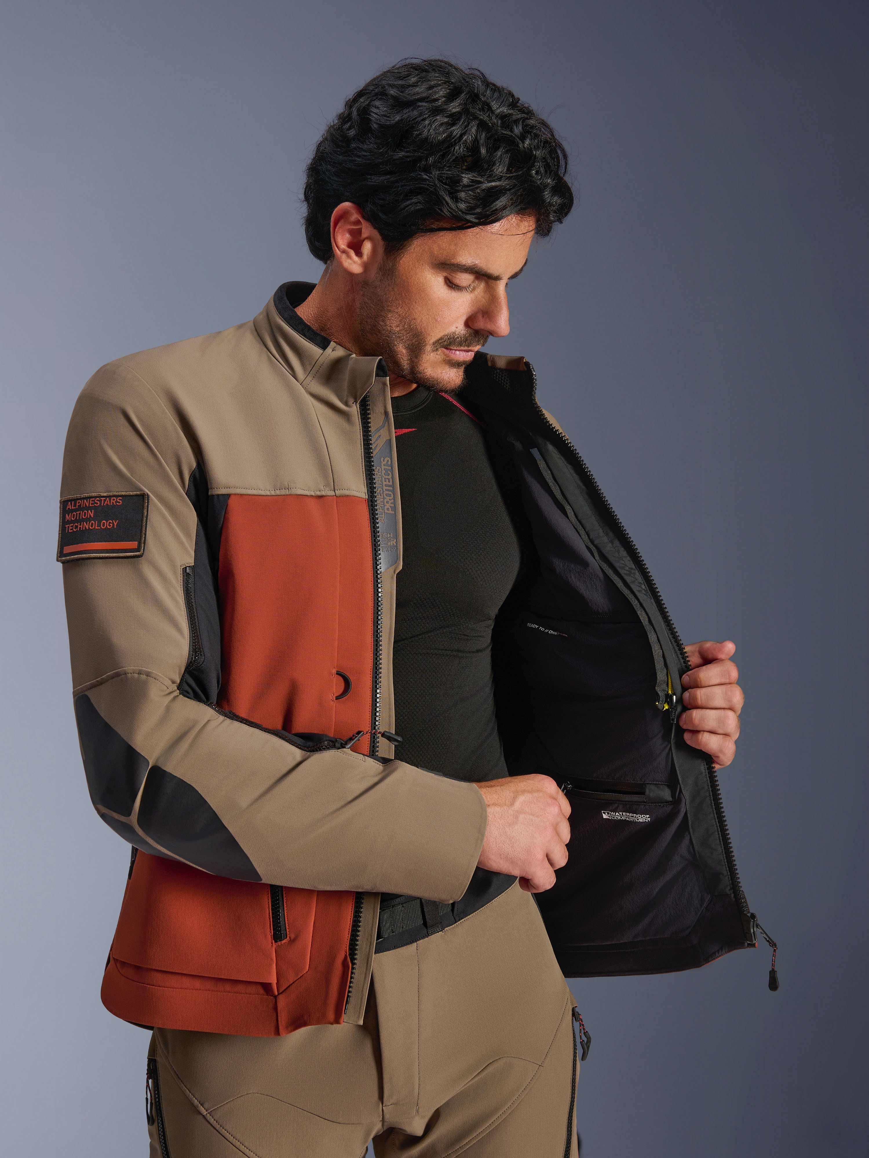 Alpinestars AMT-8 Stretch Drystar® XF Jas, Walnut Rust Black, man draagt de jas open om het interne waterdichte compartiment en de technische voering te tonen, voorzien van de 'Alpinestars Motion Technology' patch op de arm, adventure motorkleding.