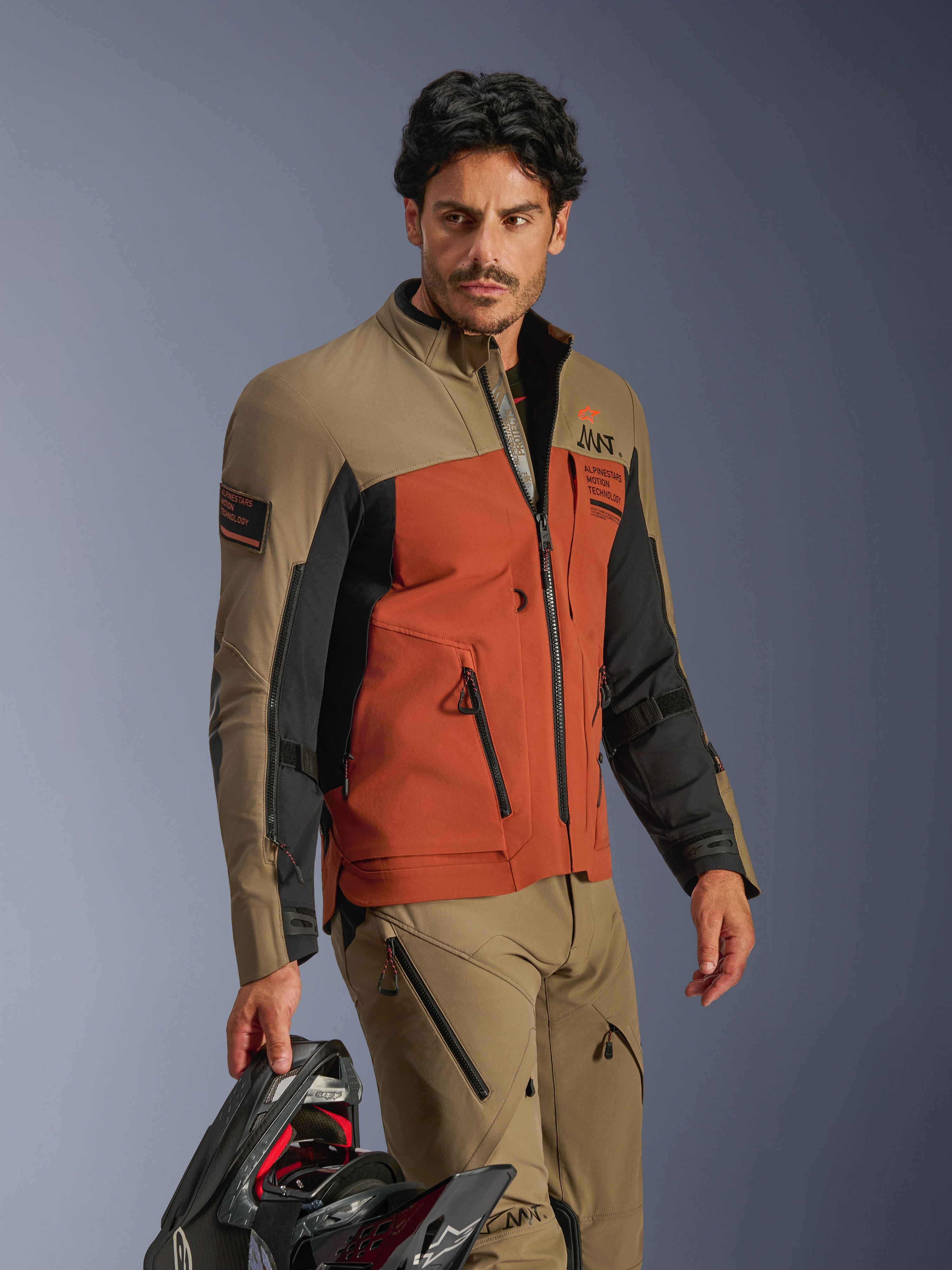 Alpinestars AMT-8 Stretch Drystar® XF Jas, Walnut Rust Black, adventure motorjas van hoogwaardig stretchmateriaal, gedragen met bijpassende walnootkleurige broek en een zwarte helm in de hand, adventure touring-uitrusting voor lange ritten