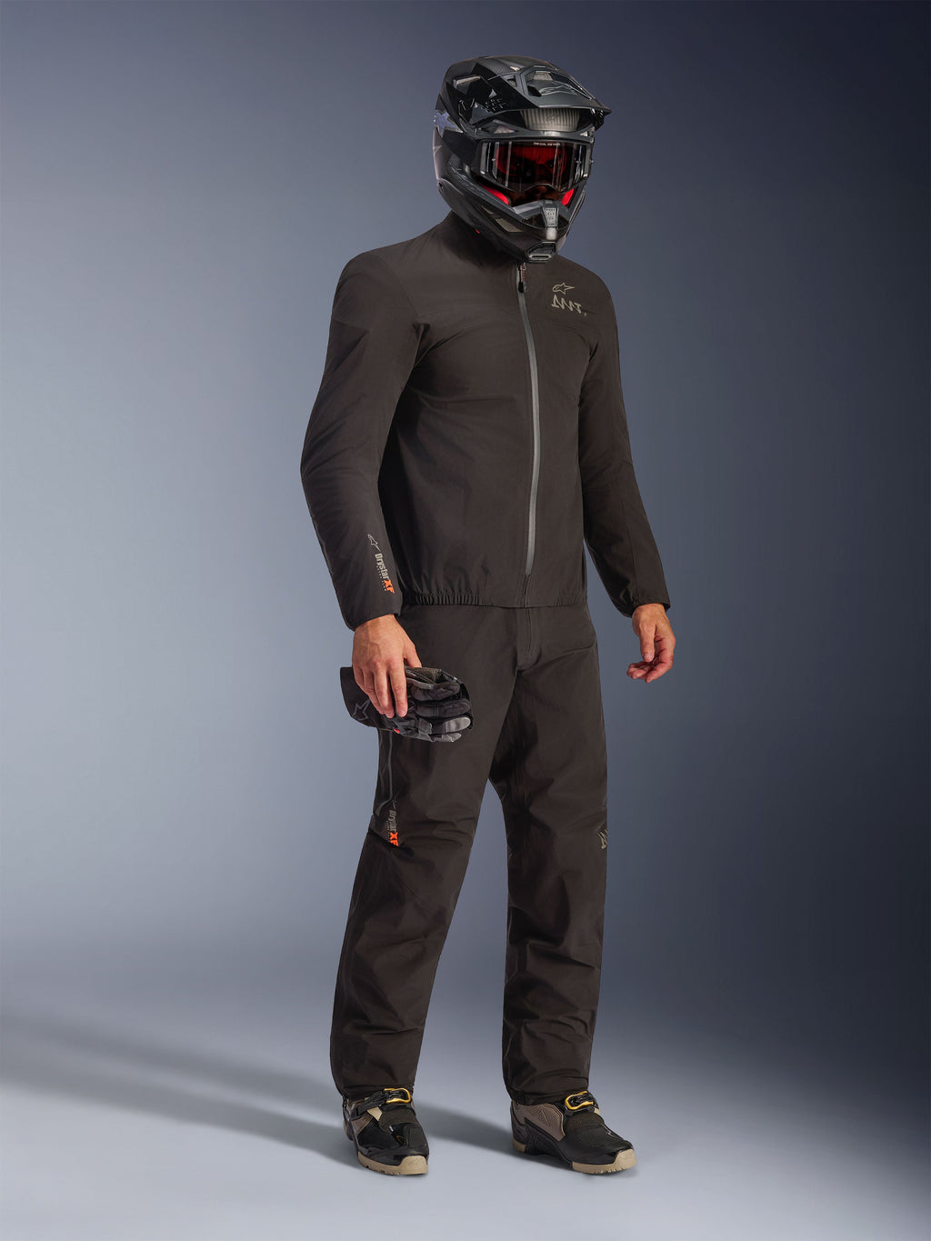 Alpinestars AMT-8 Stretch Drystar® XF Jas in zwart, gedragen als onderdeel van een complete motoroutfit met bijpassende broek en een zwarte helm, voorzien van een strak textielontwerp met oranje Drystar XF-branding op de mouw, ideaal voor adventure touring.