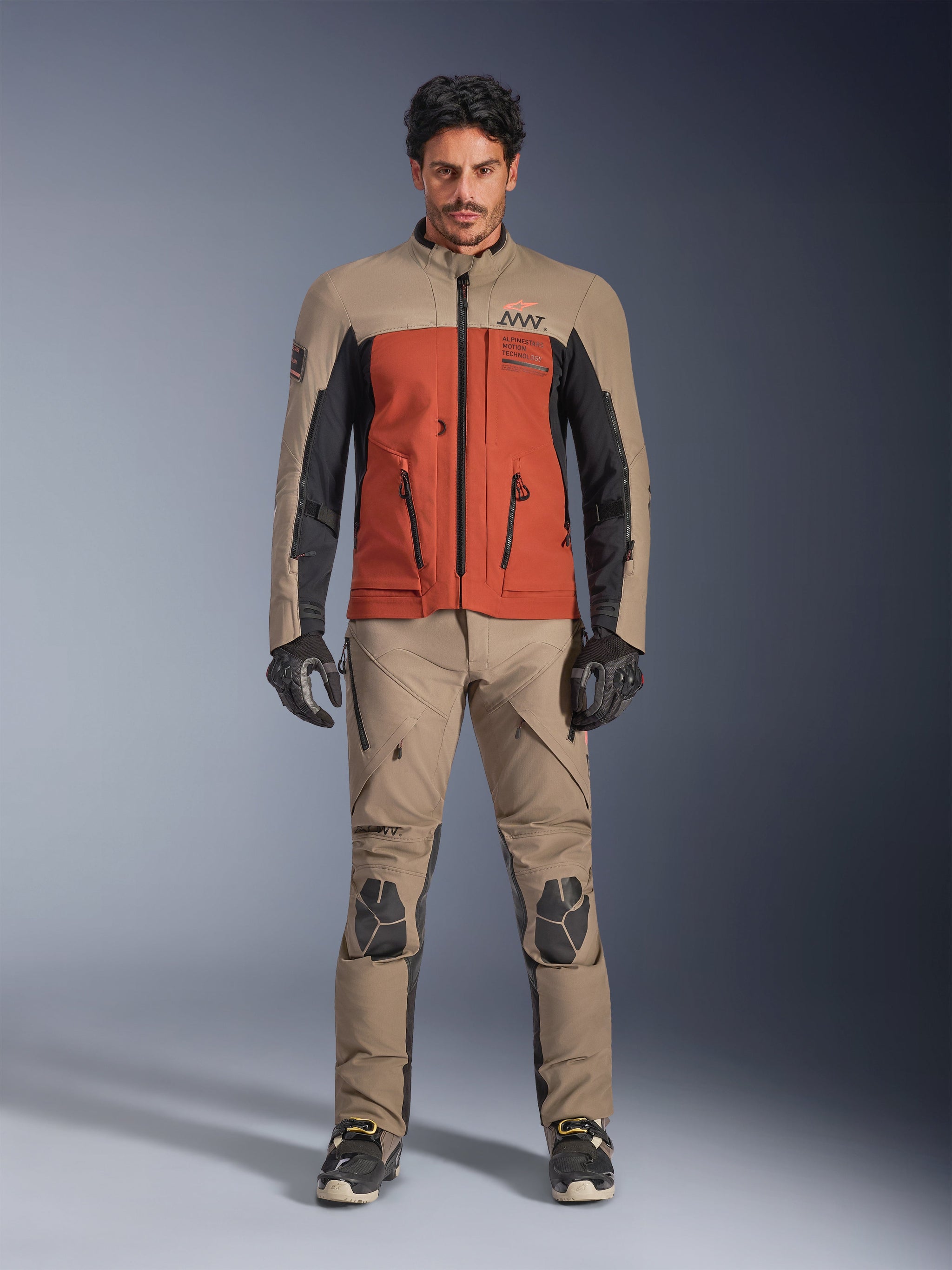 Alpinestars AMT-8 Stretch Drystar® XF Jacket, Walnut Rust Black adventure motorcycle jacket, worn by model with matching technical pants and gloves, featuring a multi-tone stretch textile design with front cargo pockets and branding, front view.