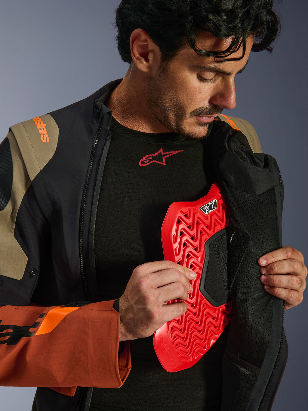 Alpinestars Halo Pro Drystar® XF gelamineerde jas in Walnut Rust Black, lifestyle-opname van een mannelijk model die de adventure-jas openritst om een rode borstprotector te onthullen, met oranje branding op de schouder en een zwarte basislaag met een rood logo.