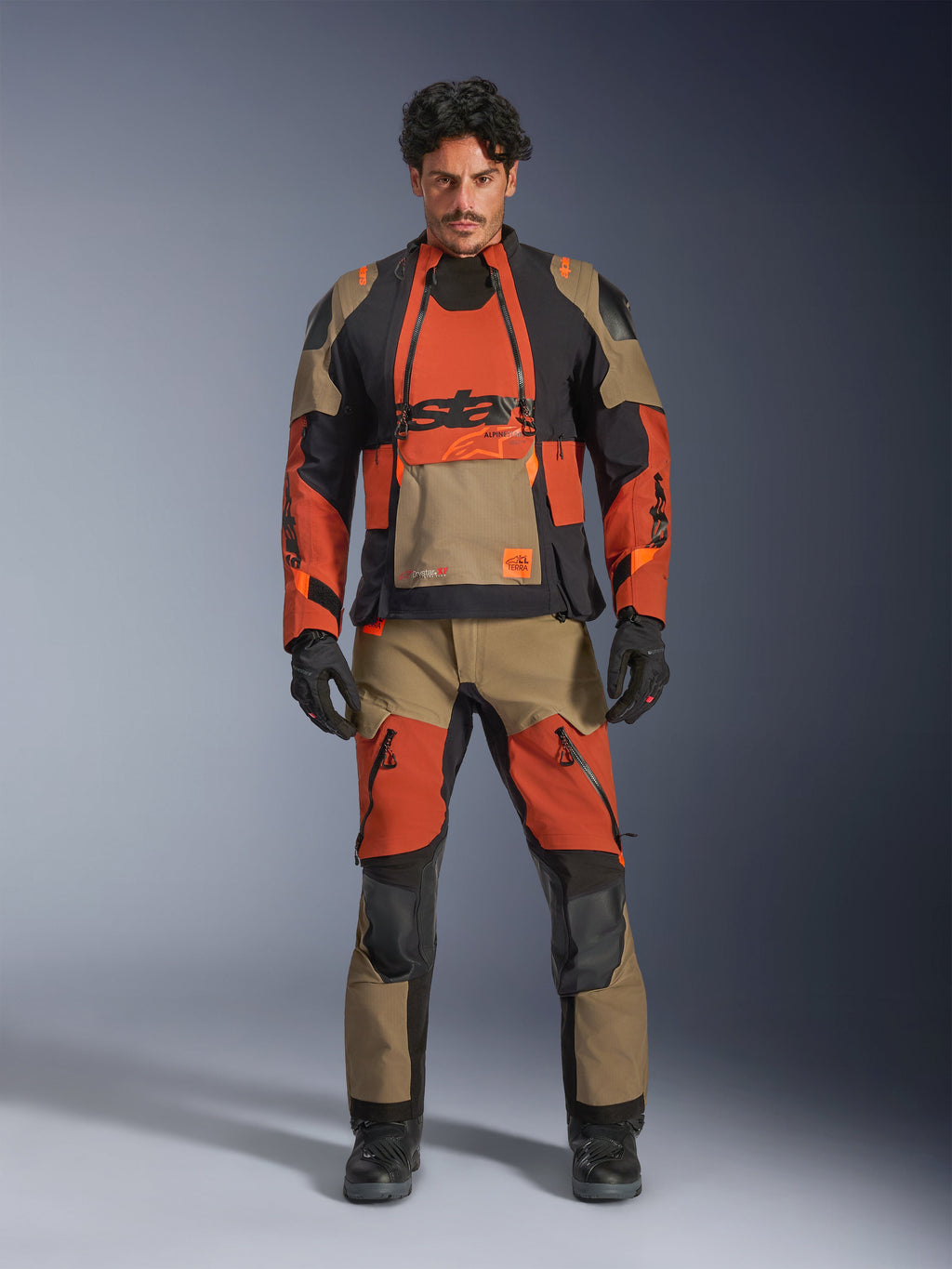 Mannelijk model draagt de Alpinestars Halo Pro Drystar® XF Gelamineerde Jas en bijpassende broek in Walnut Rust Black, modulaire adventure motoruitrusting voorzien van ventilatiepanelen aan de voorzijde en ritsen, gecombineerd met zwarte motorhandschoenen en laarzen.