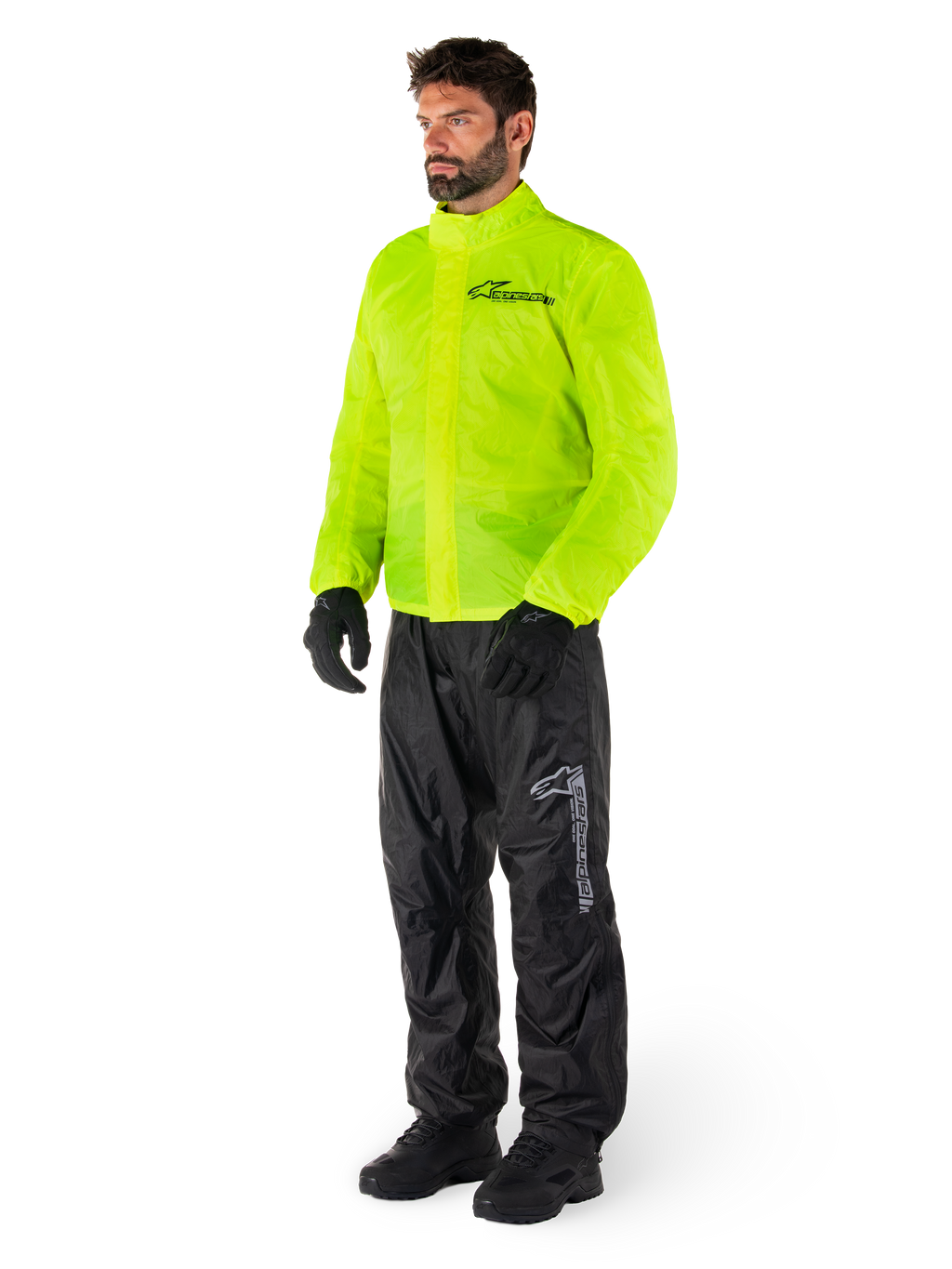 Man draagt Alpinestars Hurricane Rain V2-jas in Yellow Fluo en Hurricane Rain V2-broek in het zwart, goed zichtbare waterdichte motorregenkleding, voorzien van reflecterende logo's op de borst en het bovenbeen, complete outfit met zwarte handschoenen en laarzen.