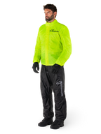 Man draagt Alpinestars Hurricane Rain V2-jas in Yellow Fluo en Hurricane Rain V2-broek in het zwart, goed zichtbare waterdichte motorregenkleding, voorzien van reflecterende logo's op de borst en het bovenbeen, complete outfit met zwarte handschoenen en laarzen.