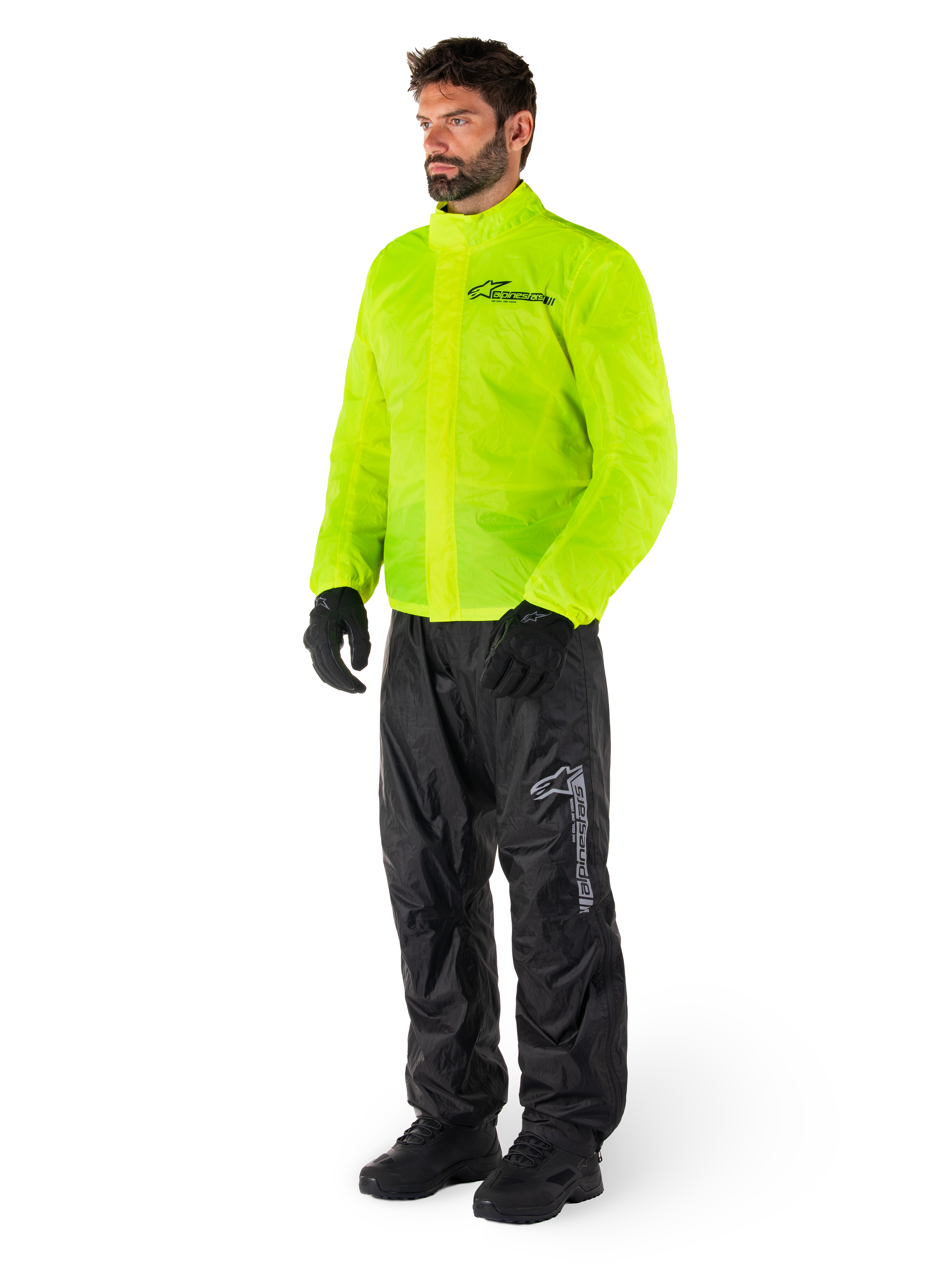 Man draagt Alpinestars Hurricane Rain V2-jas in Yellow Fluo en Hurricane Rain V2-broek in het zwart, goed zichtbare waterdichte motorregenkleding, voorzien van reflecterende logo's op de borst en het bovenbeen, complete outfit met zwarte handschoenen en laarzen.