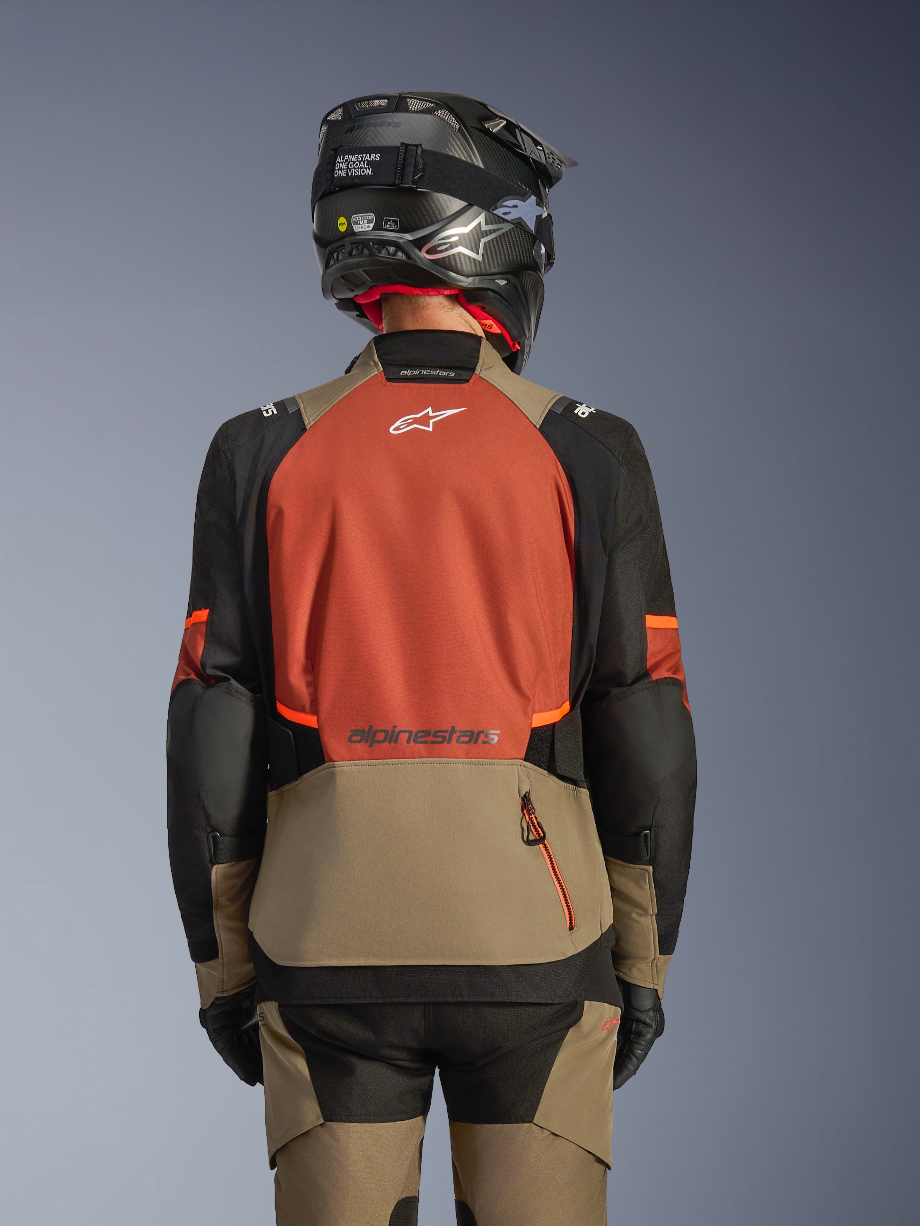 Achteraanzicht van de Alpinestars Andes V4 Drystar® jas in Walnut Rust Black, adventure touring jas met een groot roestkleurig rugpaneel met een wit logo, walnootkleurig onderste gedeelte met een verticale ritszak, zwarte mouwen met oranje accenten, gecombineerd met een bijpassende broek en een zwarte helm voor rijden in alle weersomstandigheden.