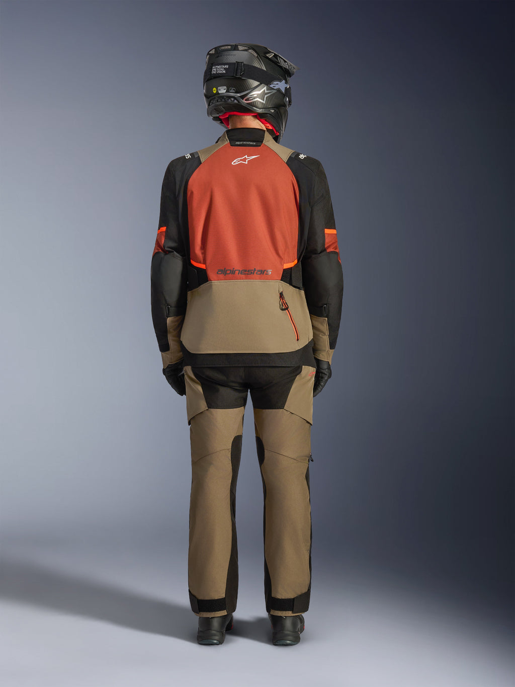 Alpinestars Andes V4 Drystar® Jas in Walnut Rust Black, achteraanzicht van een motorrijder met bijpassende walnut adventure-broek en zwarte helm, roestkleurig rugpaneel, grote opbergzak aan de achterkant en reflecterende logo's.