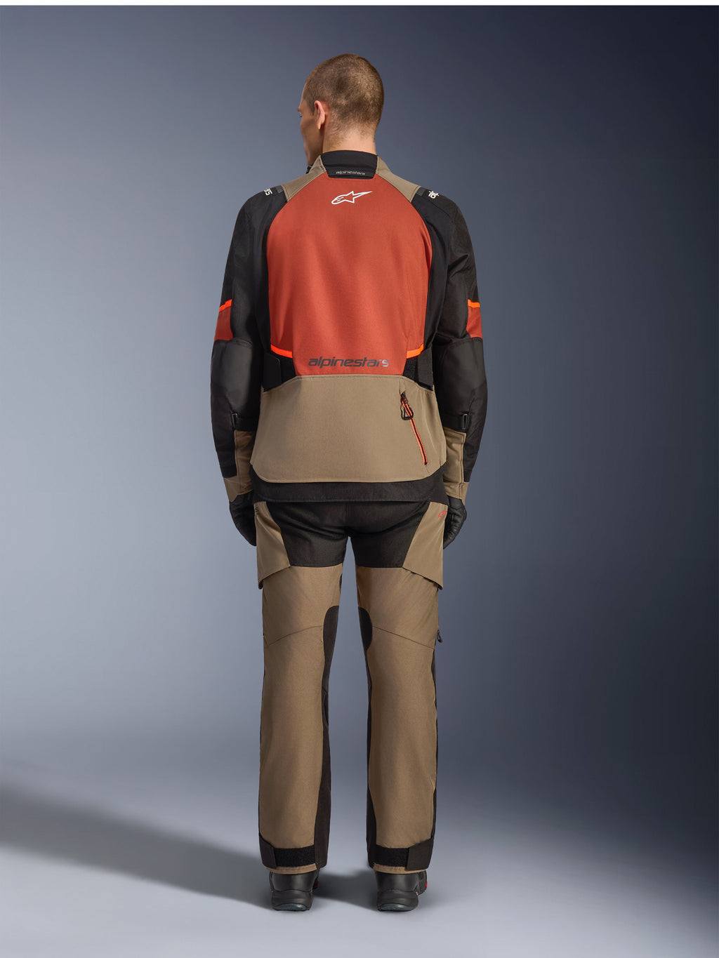 Alpinestars Andes V4 Drystar® Jas, Walnut Rust Black, achteraanzicht van touringmotorjas met roestkleurig bovenpaneel op de rug en walnootkleurig onderste gedeelte, gedragen met bijpassende adventure-broek en zwarte laarzen