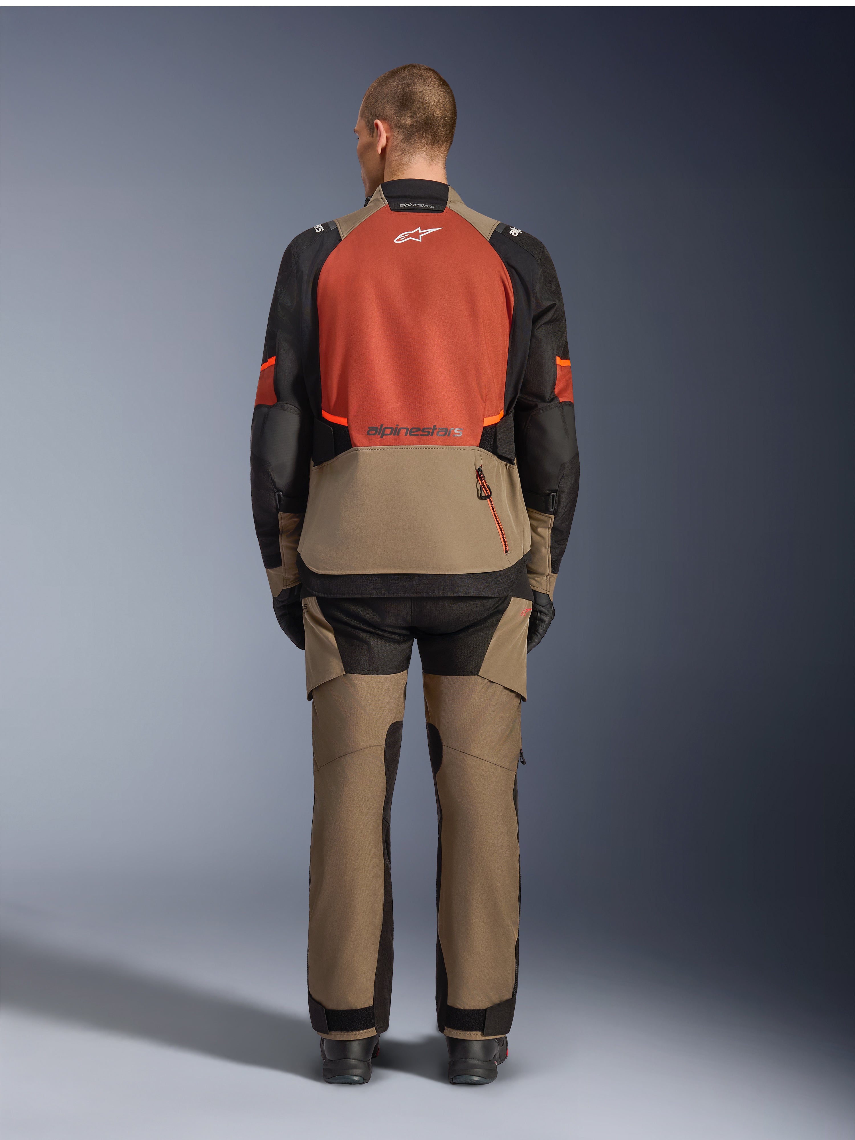 Alpinestars Andes V4 Drystar® Jas, Walnut Rust Black, achteraanzicht van touringmotorjas met roestkleurig bovenpaneel op de rug en walnootkleurig onderste gedeelte, gedragen met bijpassende adventure-broek en zwarte laarzen