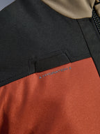 Close-up detail van de Alpinestars Andes V4 Drystar® Jas in Walnut Rust Black, met de nadruk op de geïntegreerde lus voor de hydratatieslang op de borst en de duurzame ripstop textielconstructie.