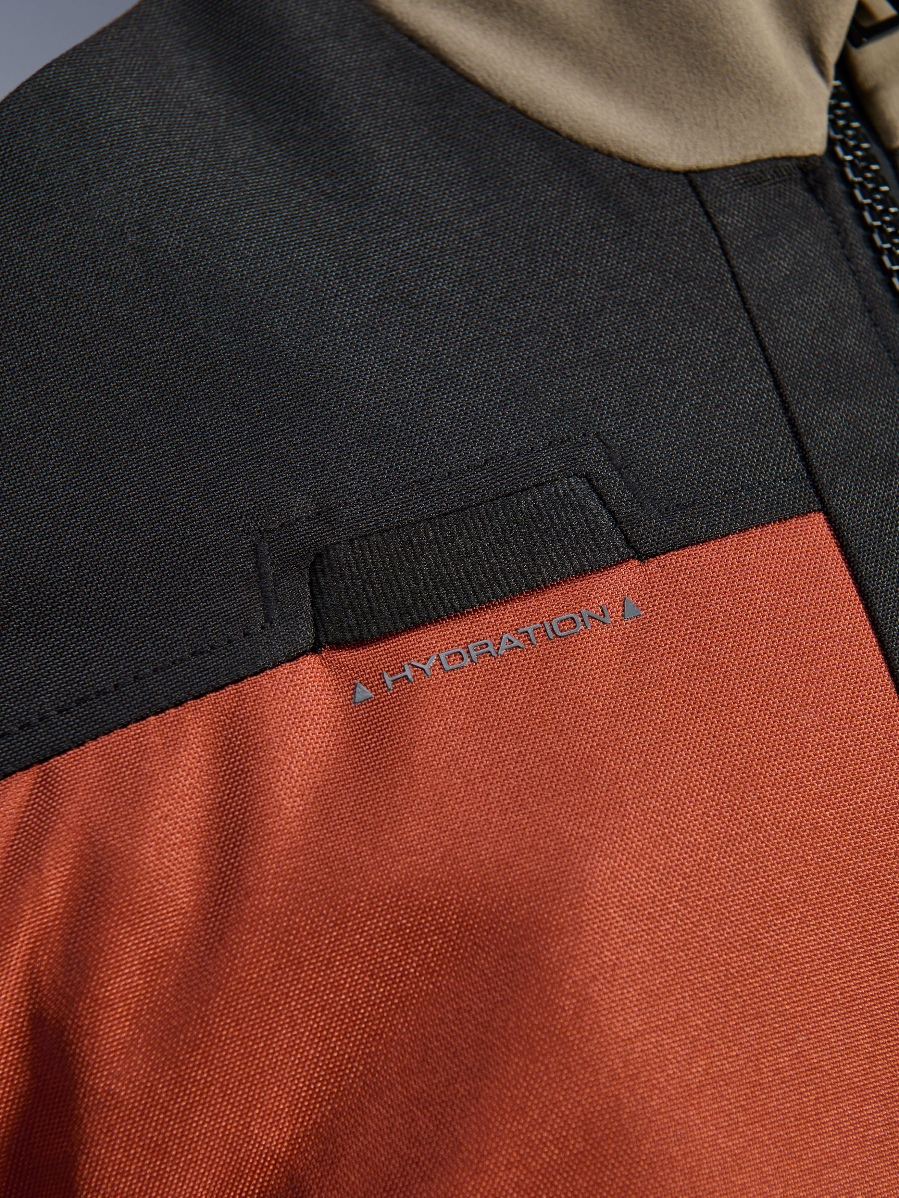 Close-up detail van de Alpinestars Andes V4 Drystar® Jas in Walnut Rust Black, met de nadruk op de geïntegreerde lus voor de hydratatieslang op de borst en de duurzame ripstop textielconstructie.