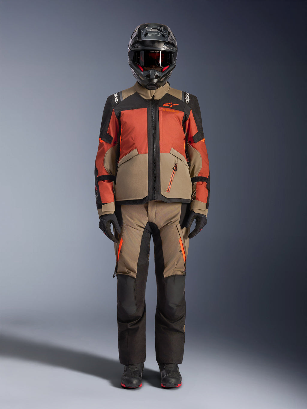 Adventure rijder die de Alpinestars Andes V4 Drystar® Jas in Walnut Rust Black draagt, gecombineerd met een bijpassende adventure broek, een zwarte helm met rode accenten en motorbril, en zwarte handschoenen, wat een complete all-weather touring-outfit toont.