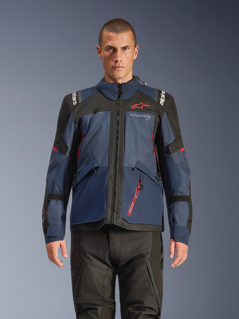 Alpinestars Andes V4 Drystar® jas, adventure motorjas, Night Navy en Mood Indigo met zwarte panelen, rood Alpinestars-logo op de borst en rood ritsdetail, duurzame textielconstructie voor toerritten onder alle weersomstandigheden, gedragen door model met zwarte motorbroek.