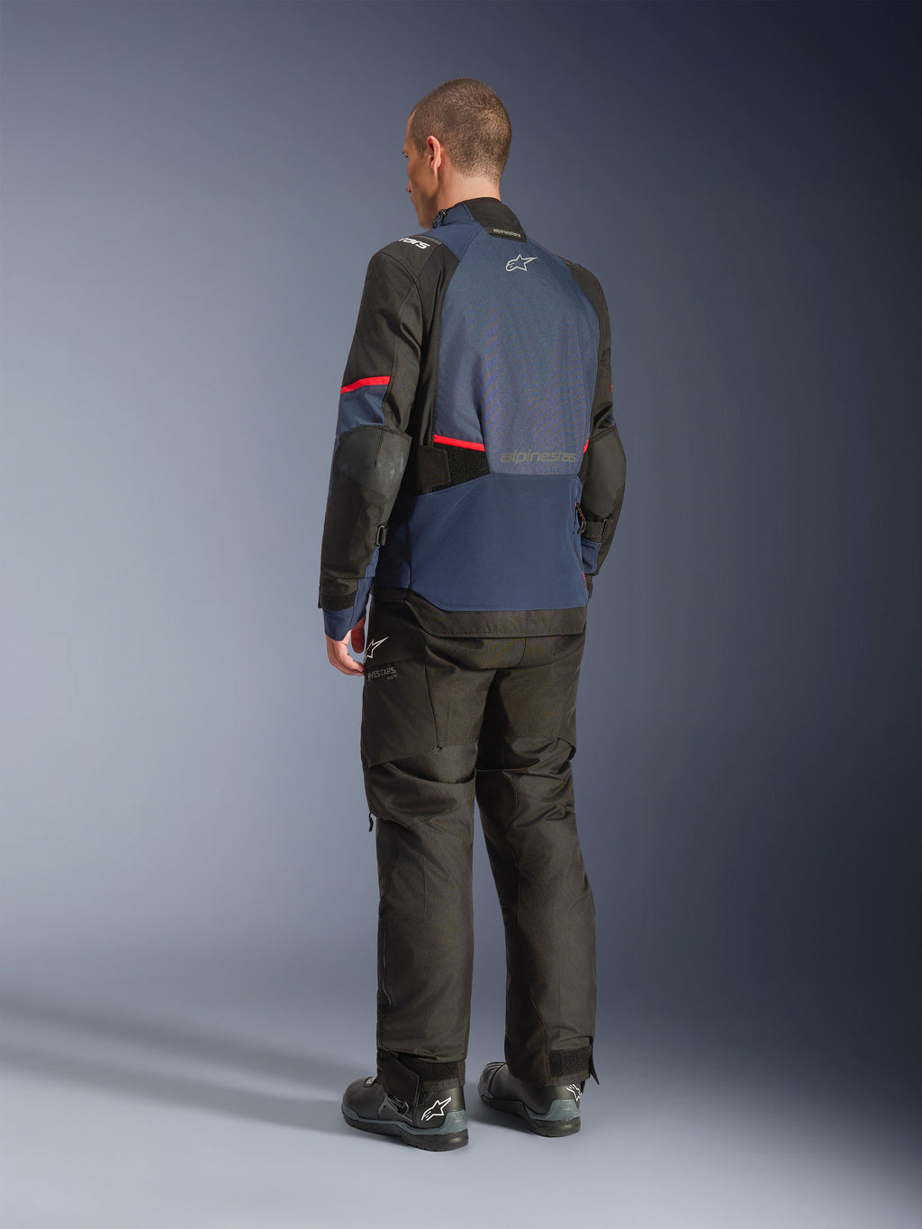 Alpinestars Andes V4 Drystar® Jas, Night Navy en Mood Indigo met zwarte panelen en rode accenten, driekwart achteraanzicht, gedragen met bijpassende zwarte adventure motorbroek en laarzen, technische textielconstructie met Alpinestars-logo op de bovenrug, verlengde achterzoom voor extra bedekking.