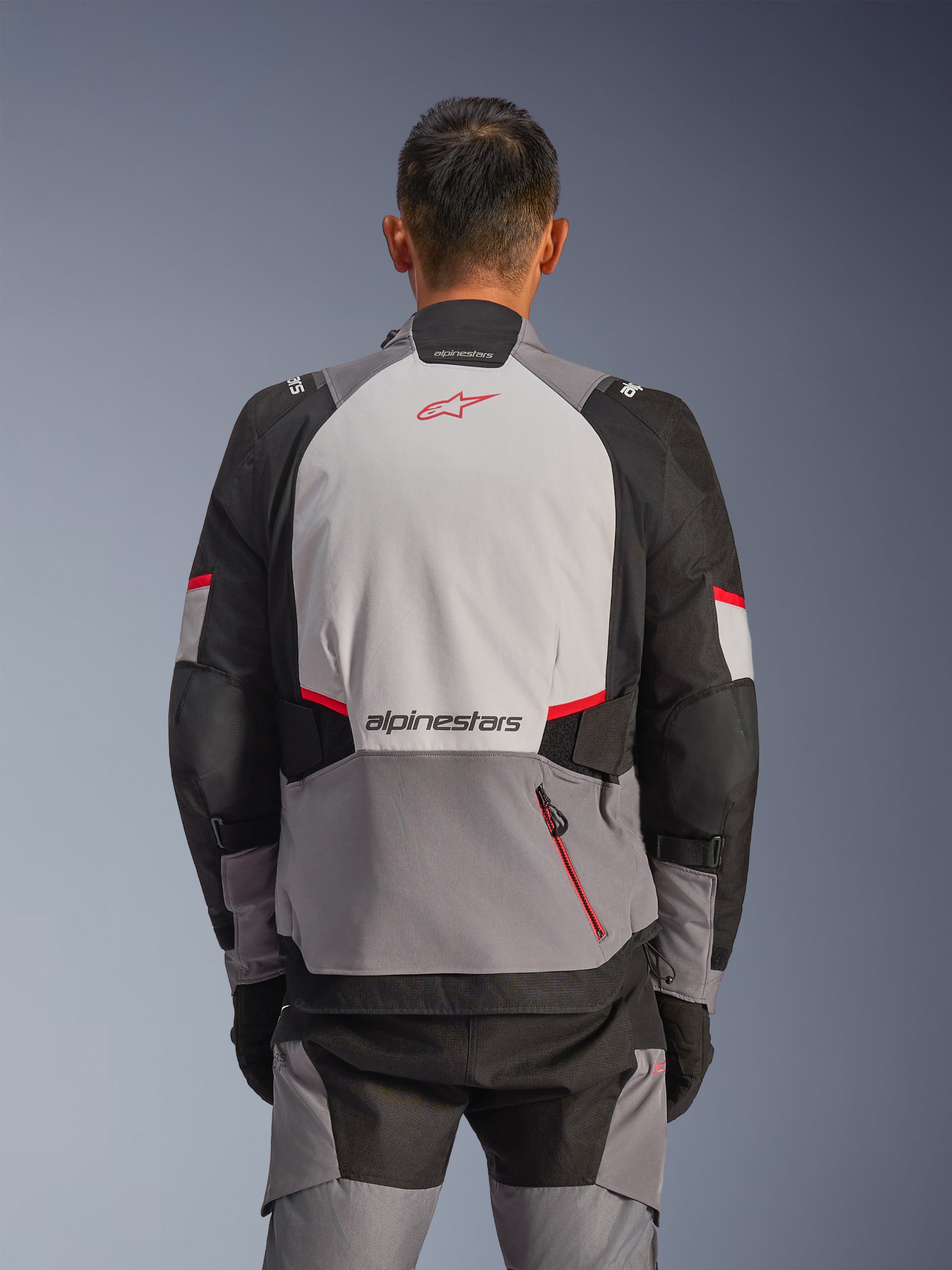 Alpinestars Andes V4 Drystar® jas, achteraanzicht, donkergrijs, ijsgrijs, zwart en felrood, voorzien van een rood Astar-logo en Alpinestars-woordmerk op de rug, versterkte panelen en een zak met rode rits, adventure motorrijuitrusting