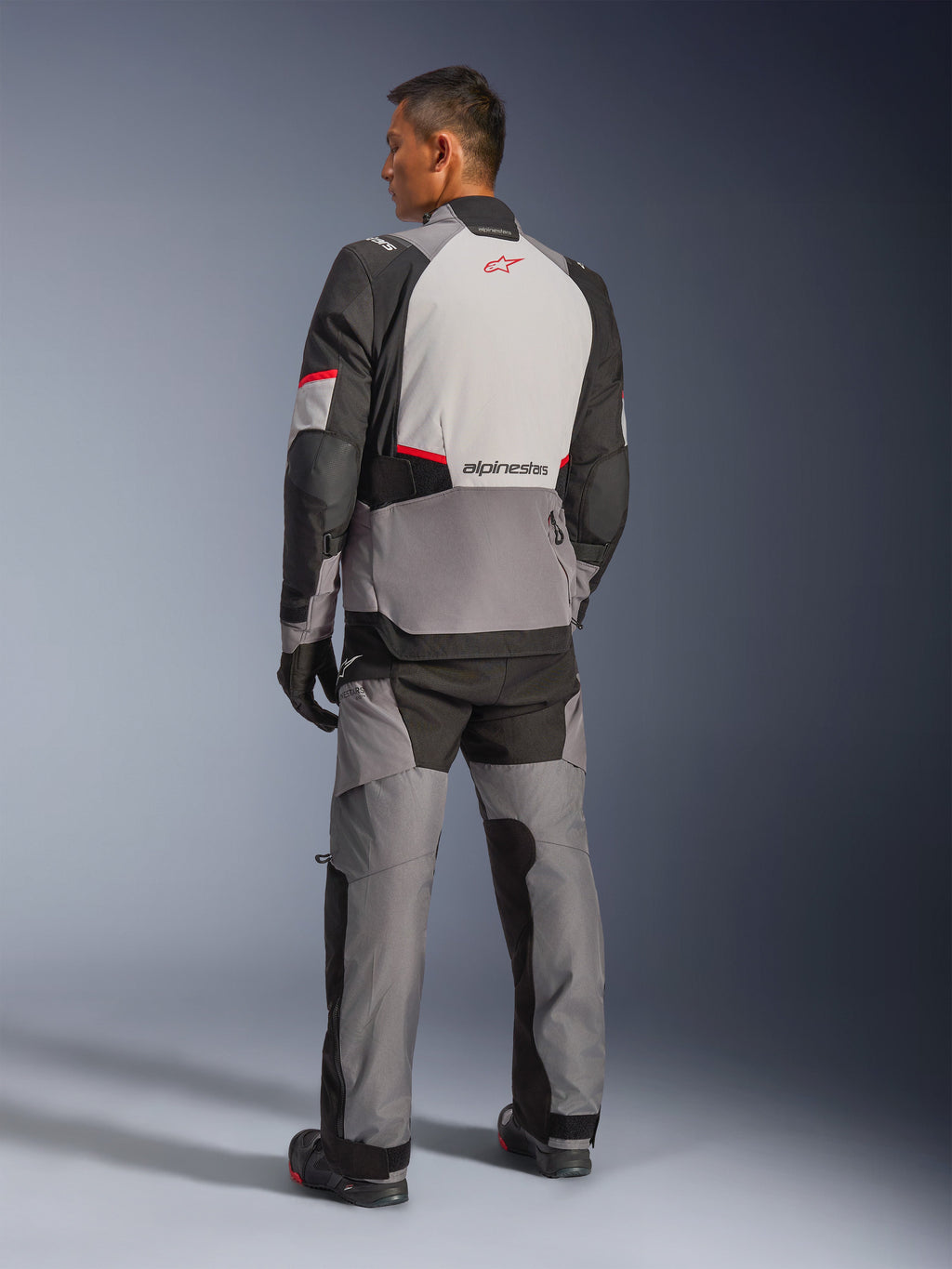 Alpinestars Andes V4 Drystar® Jas, donkergrijs en Ice Gray met zwarte en felrode accenten, achteraanzicht met lichtgrijs paneel en rood sterlogo, gecombineerd met bijpassende adventure broek en zwarte handschoenen, technische motoruitrusting voor alle weersomstandigheden.
