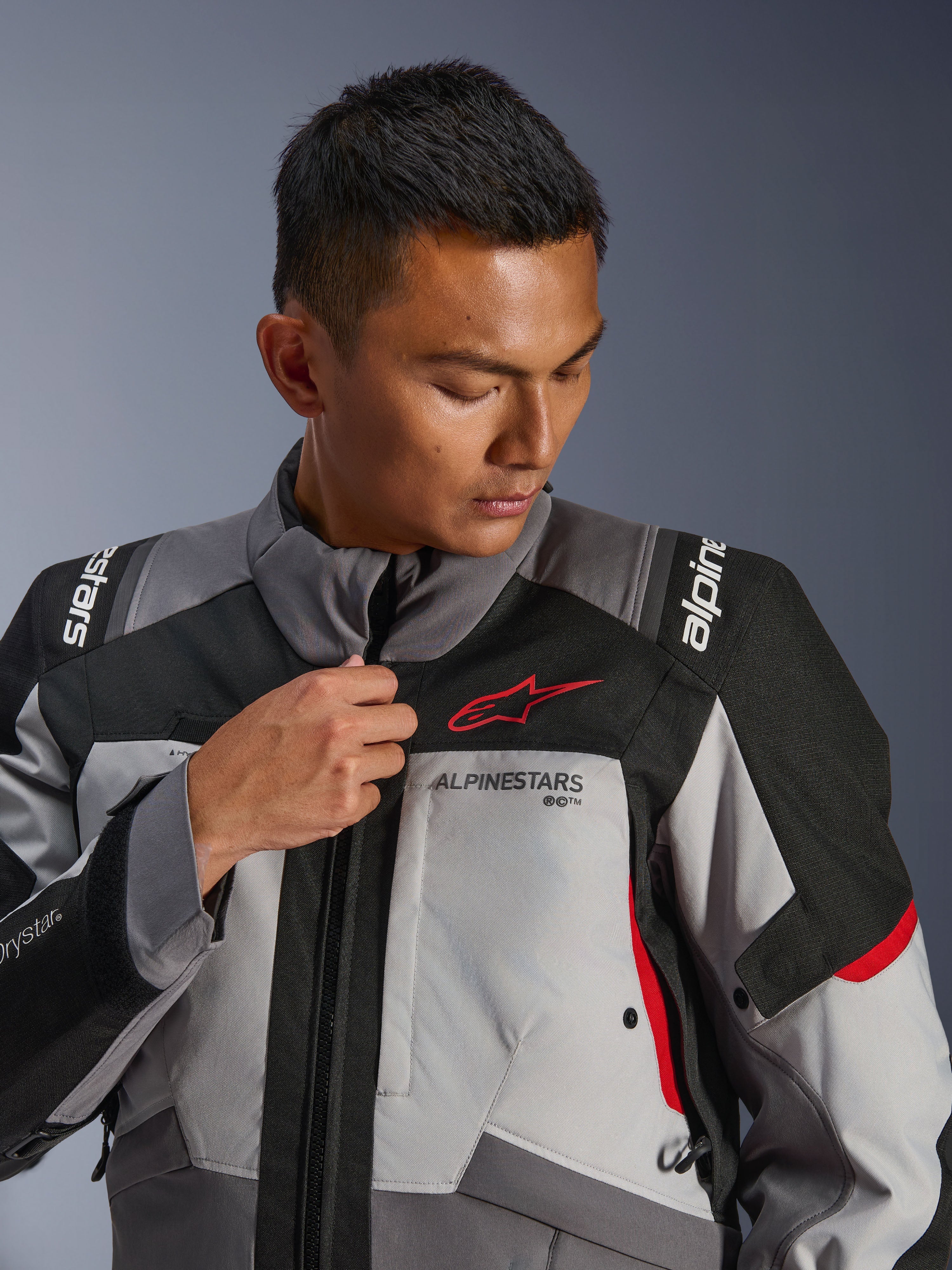 Model draagt Alpinestars Andes V4 Drystar® Jacket, donkergrijs en ijsgrijs met zwarte en felrode accenten, verstelt de rits aan de voorzijde, met het rode Astars-logo en Drystar®-branding, adventure touring-stijl