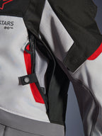 Detailopname van de Alpinestars Andes V4 Drystar® Jacket in donkergrijs, ijsgrijs en felrood, met een ventilatieopening met ritssluiting op de borst, mesh-voering en een verstelbandje met drukknoop.