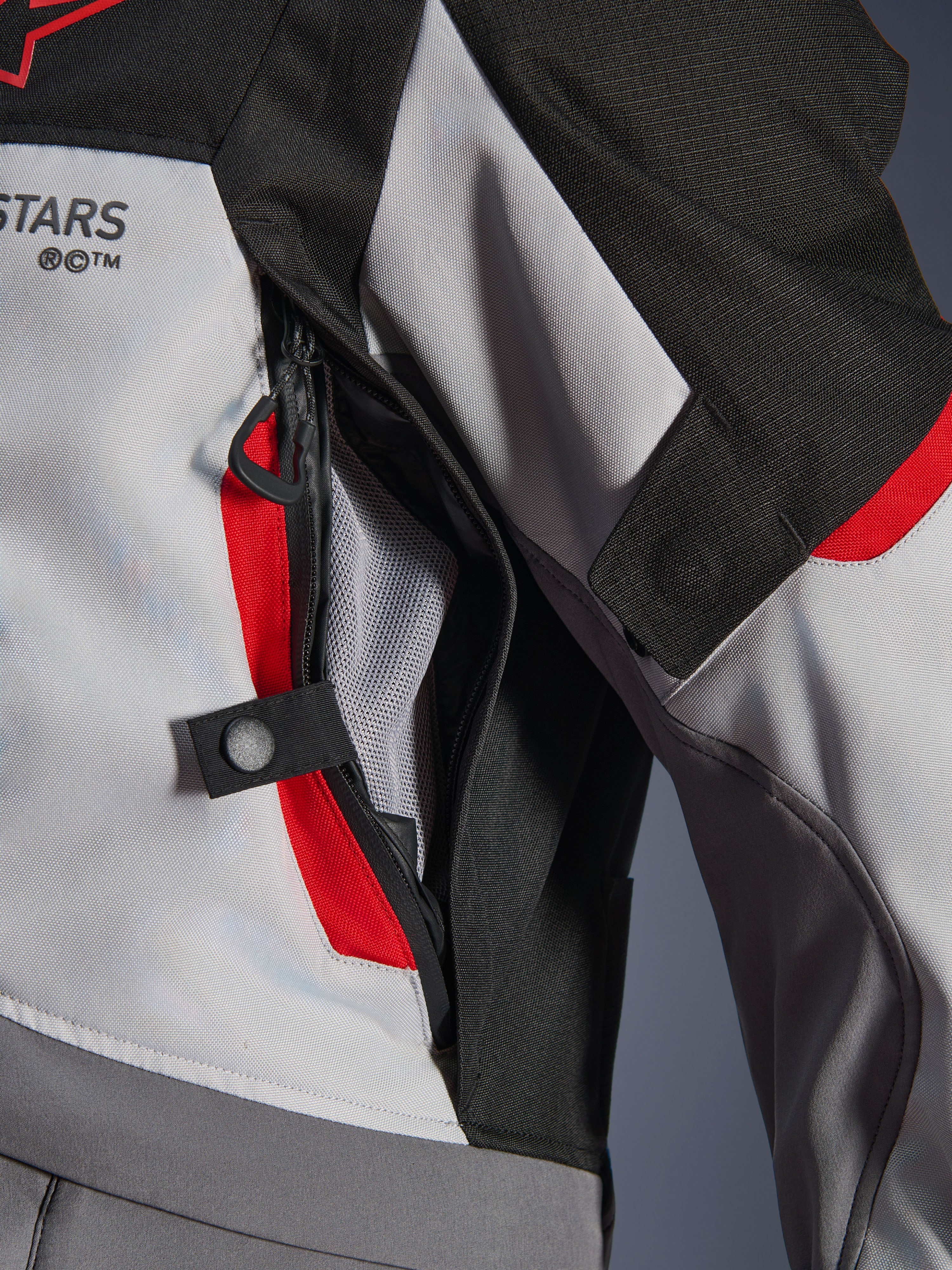 Detailopname van de Alpinestars Andes V4 Drystar® Jacket in donkergrijs, ijsgrijs en felrood, met een ventilatieopening met ritssluiting op de borst, mesh-voering en een verstelbandje met drukknoop.