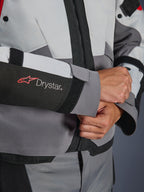 Close-up detail van de mouw van de Alpinestars Andes V4 Drystar® jas, grijs en zwart textiel, voorzien van een rood en wit Drystar-logo op de onderarm, verstelbare klittenbandsluiting bij de pols, hoogwaardige waterdichte motorkleding