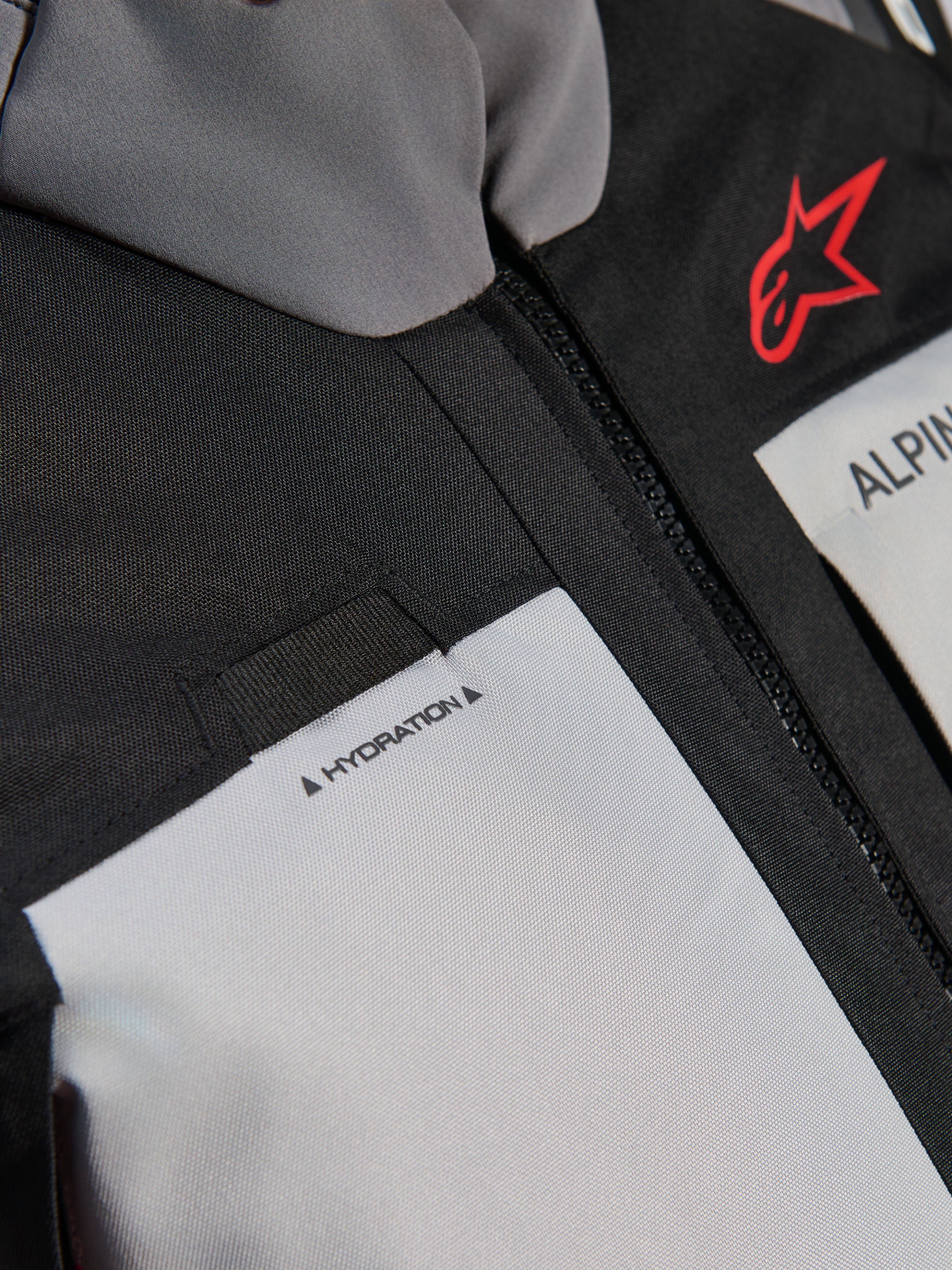 Alpinestars Andes V4 Drystar® jas, close-up detail van de borst, donkergrijze en Ice Gray panelen met zwarte textielversterkingen, voorzien van een rood Alpinestars-logo en een lus voor de hydratatieslang met 'HYDRATION'-label