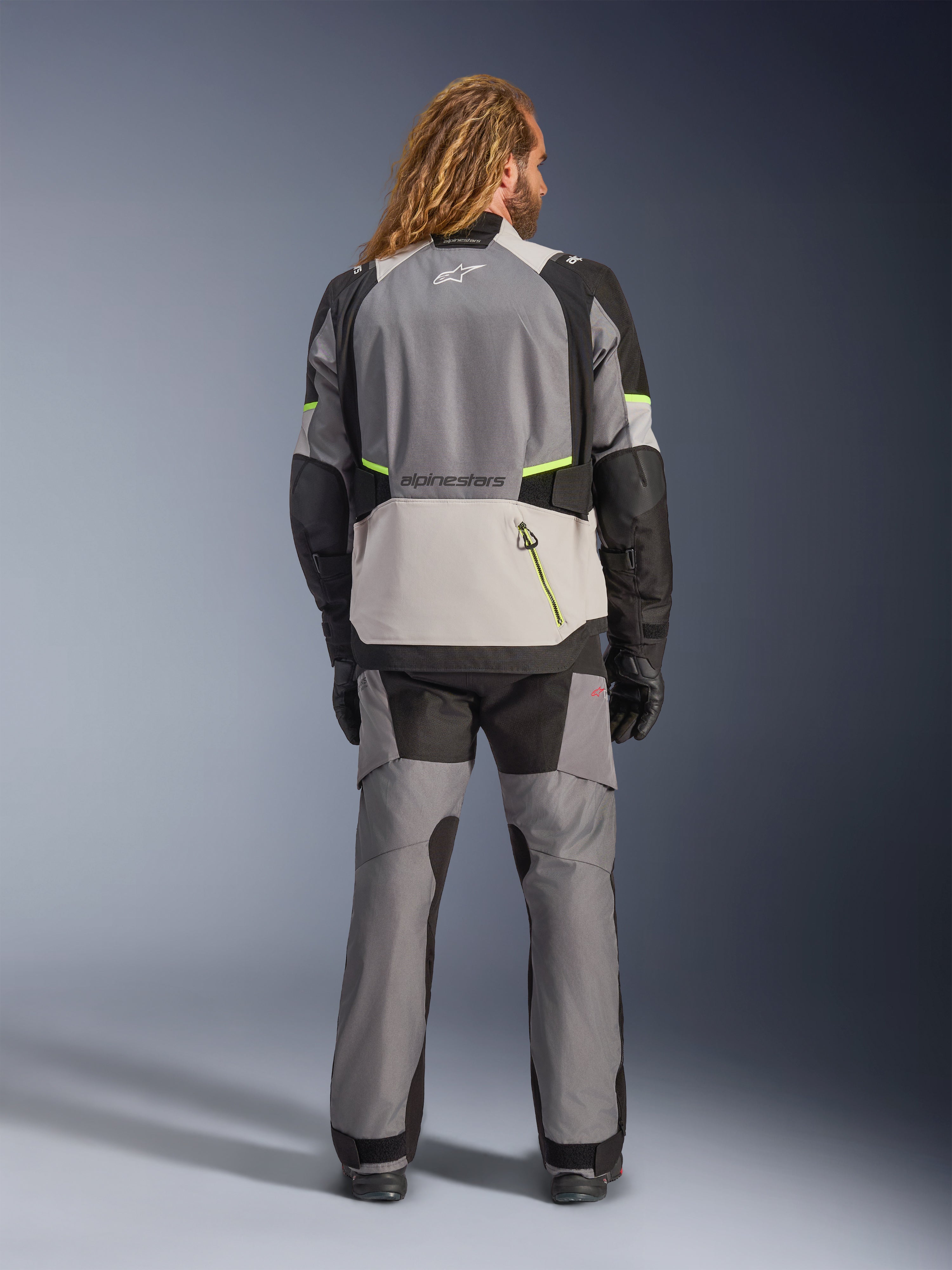 Alpinestars Andes V4 Drystar® Jas, adventure motorjas, donkergrijs en ijsgrijs met zwarte en fluo gele accenten, achteraanzicht gedragen met bijpassende broek en zwarte handschoenen, voorzien van een grote achterzak met gele rits en verstevigde schouder- en elleboogpanelen