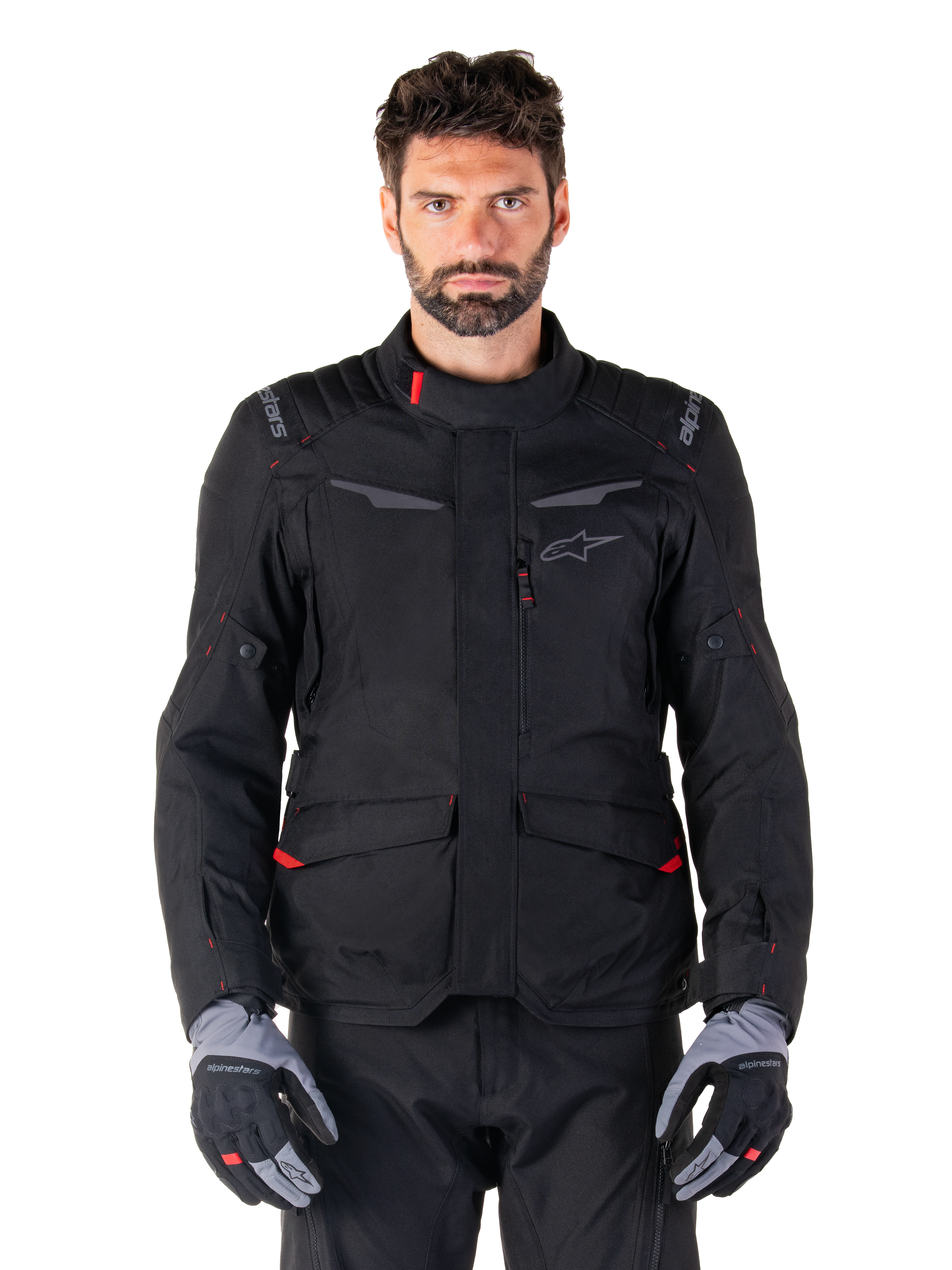 Alpinestars ST-1 Waterproof Jacket, motorjas voor adventure touring, zwart met rode accenten, hoge kraag, meerdere zakken, duurzaam getextureerd materiaal, gedragen met zwarte broek en grijze handschoenen
