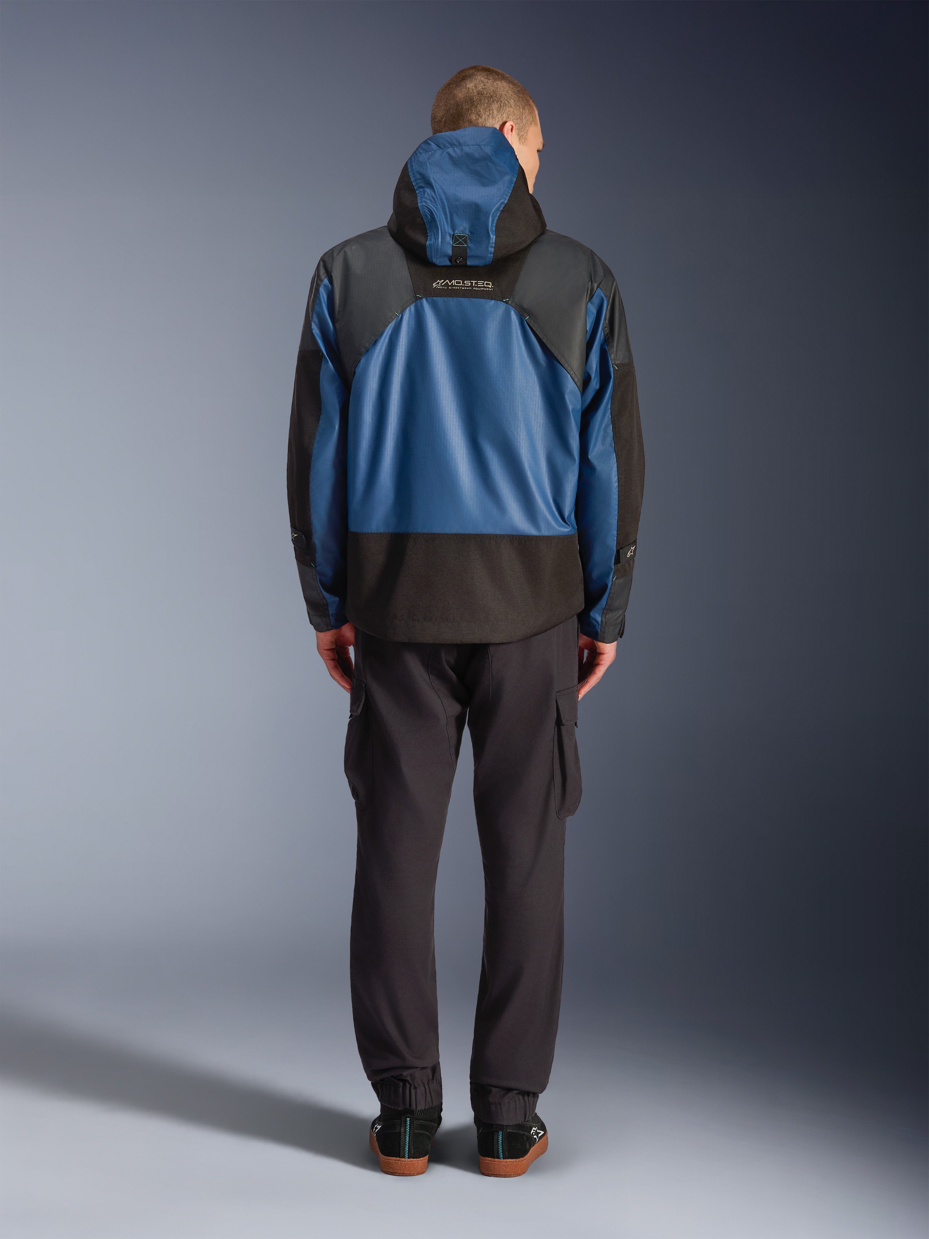 Alpinestars Mospher Waterproof jas in zwart en Dark Petrol, achteraanzicht op model, gecombineerd met een zwarte cargobroek, voorzien van tactische ripstop-constructie en MO.ST.EQ.-branding op de rug