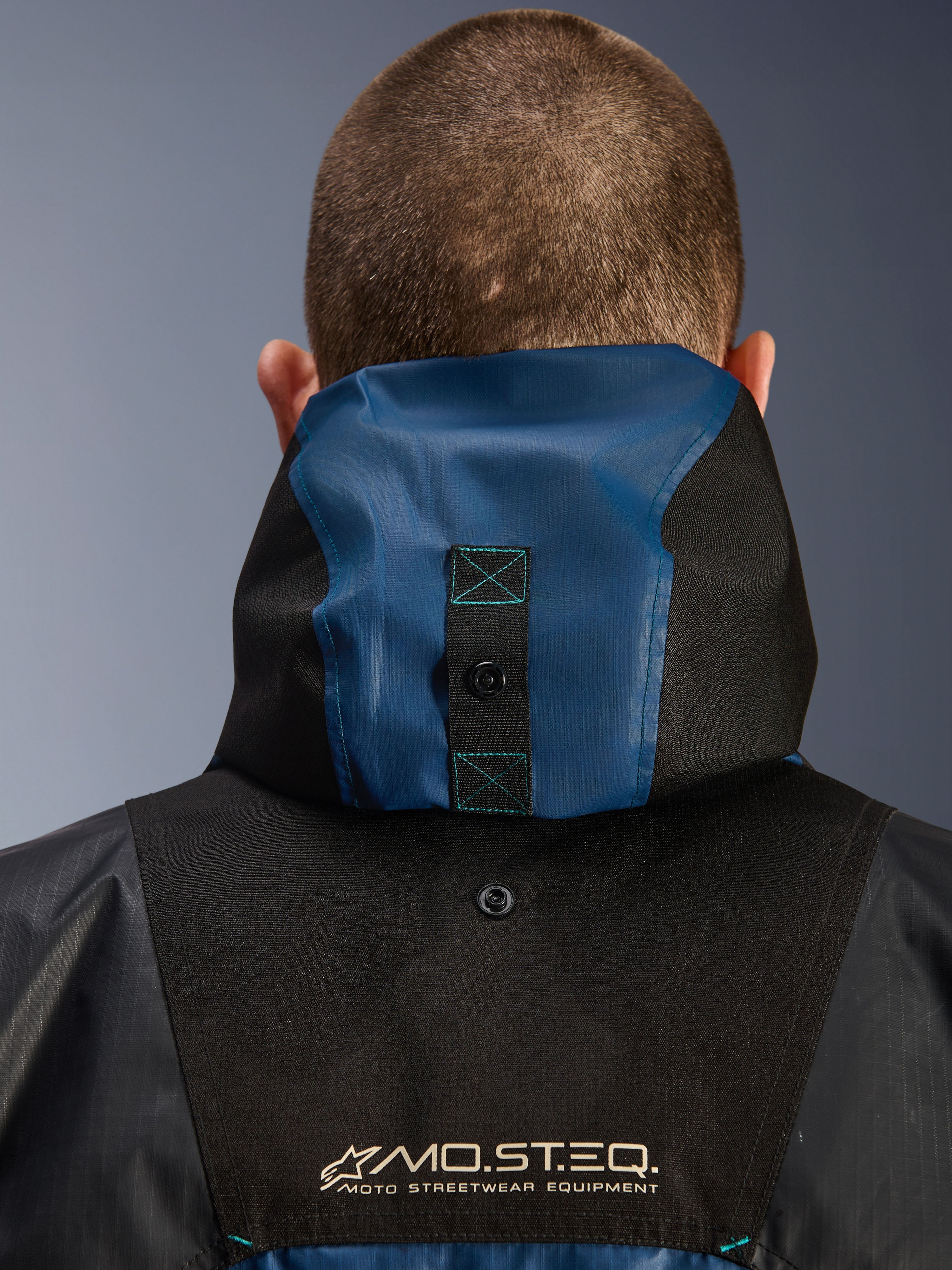 Alpinestars Mospher Waterproof Jas, detail van de achterkant met het capuchonverankeringssysteem, zwart en Dark Petrol blauw, voorzien van MO.ST.EQ. Moto Streetwear Equipment-branding, duurzaam ripstop textielmateriaal, technische motoruitrusting voor woon-werkverkeer