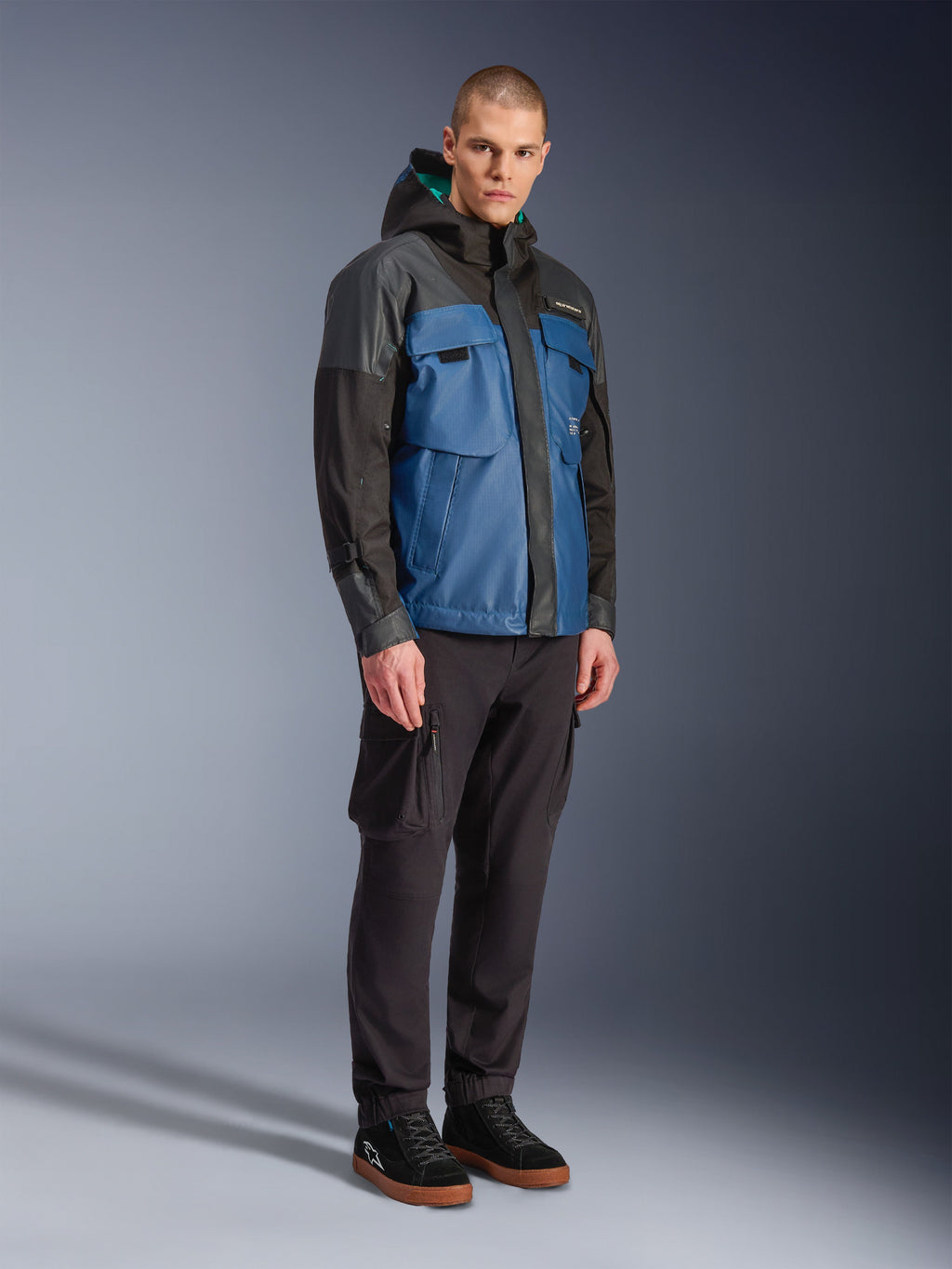 Alpinestars Mospher Waterproof Jacket, zwart en Dark Petrol blauw, gedragen met een zwarte cargobroek en zwarte sneakers, stedelijke forenzenstijl met functionele zakken en capuchon