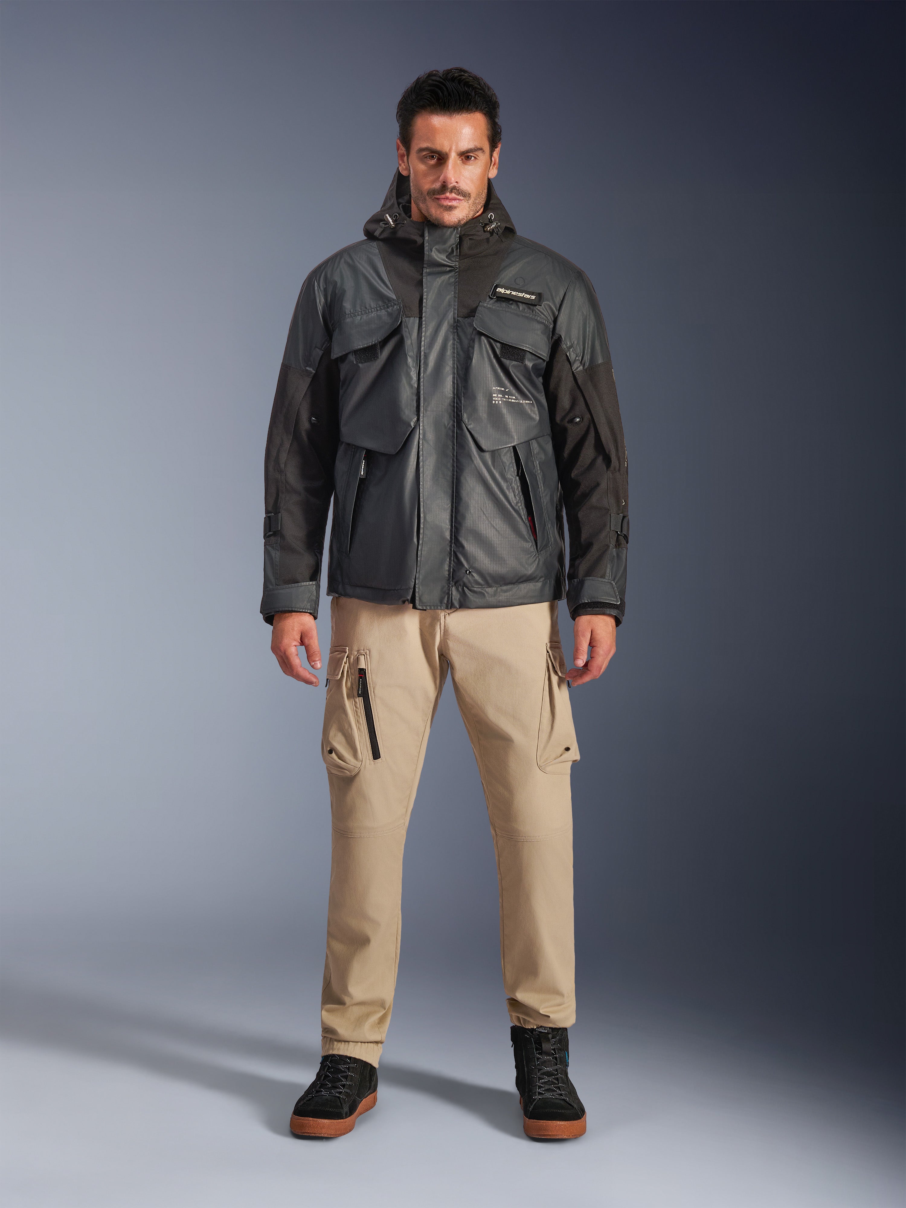 Mannelijk model in de Alpinestars Mospher Waterproof Jacket in Black, urban eigentijdse motorkleding, tactisch ontwerp met meerdere functionele zakken en capuchon, gecombineerd met een beige cargobroek en zwarte schoenen, full-body studioportret