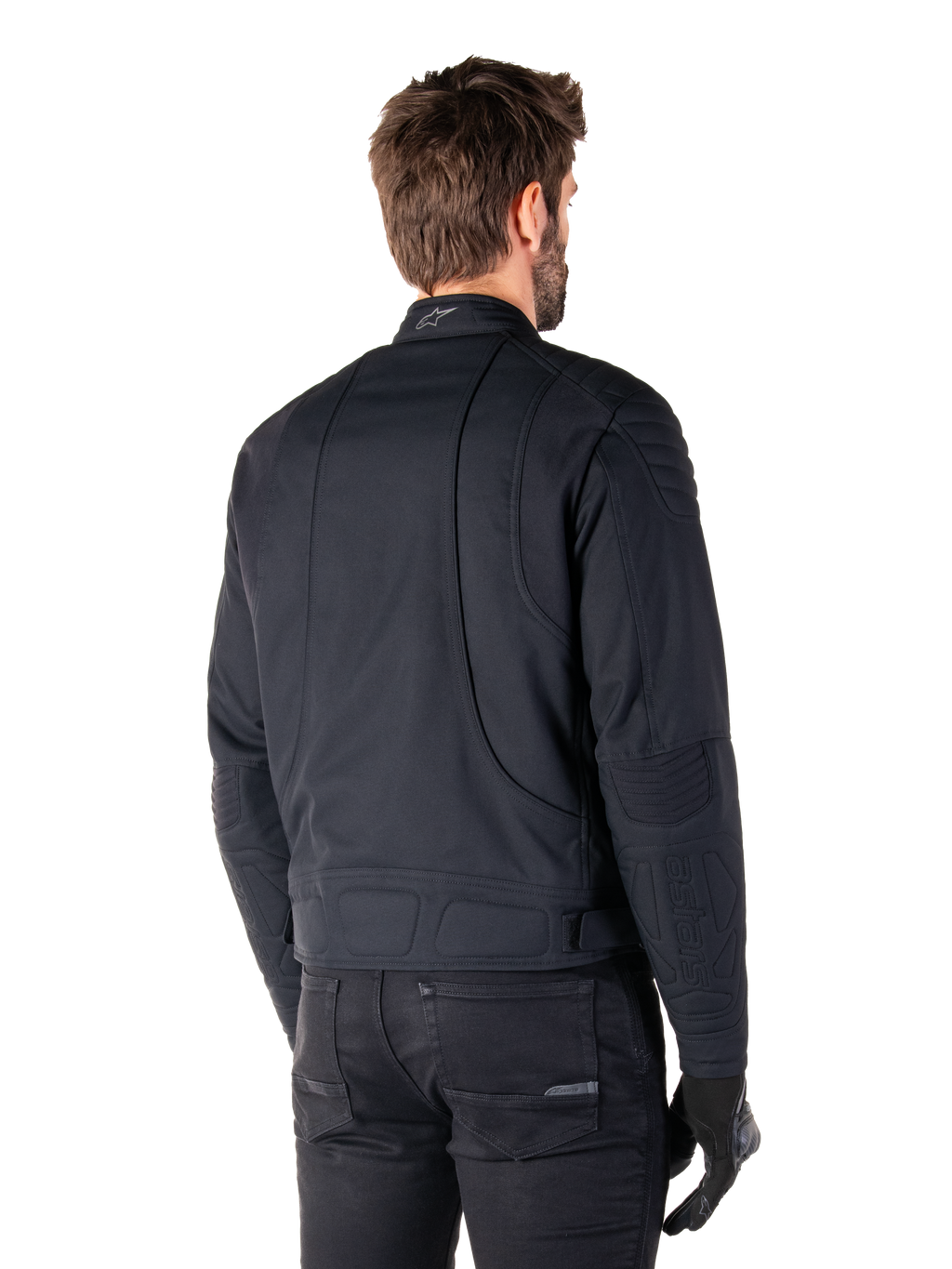 Alpinestars Clayton WR jas in het zwart, driekwart achteraanzicht op een mannelijk model, urban motorjas met gewatteerde vulling op de schouders en ellebogen, technische stiklijnen en reflecterend logo op de kraag, gecombineerd met een zwarte broek en handschoenen.