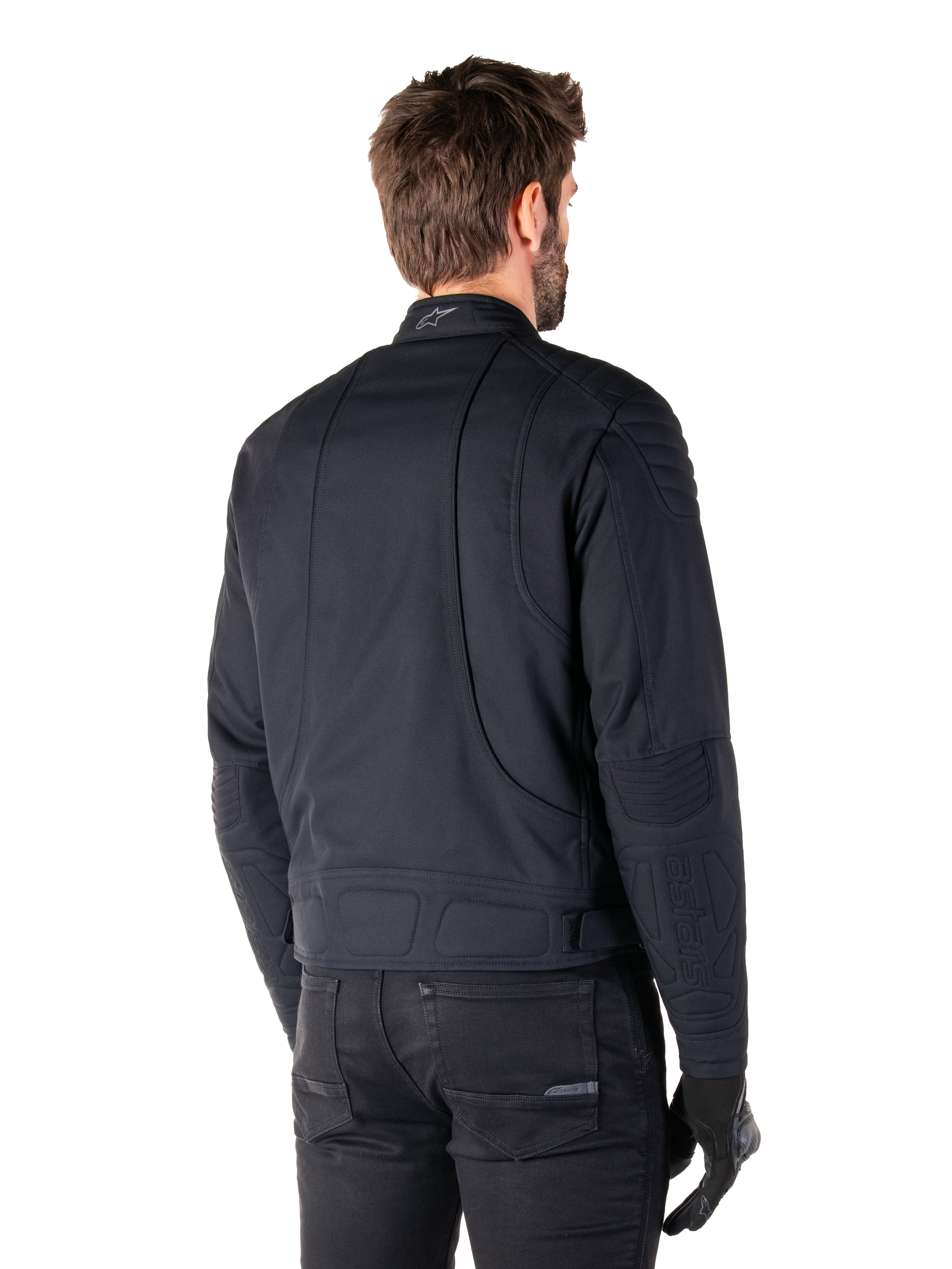 Alpinestars Clayton WR jas in het zwart, driekwart achteraanzicht op een mannelijk model, urban motorjas met gewatteerde vulling op de schouders en ellebogen, technische stiklijnen en reflecterend logo op de kraag, gecombineerd met een zwarte broek en handschoenen.