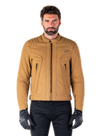 Alpinestars Clayton WR jas, urban motorjas, Utility Brown Light Gold, waterbestendig textiel met gewatteerde schouders, hoge kraag, zwarte ritsdetails en logo op de borst, gedragen met zwarte beschermende handschoenen en broek
