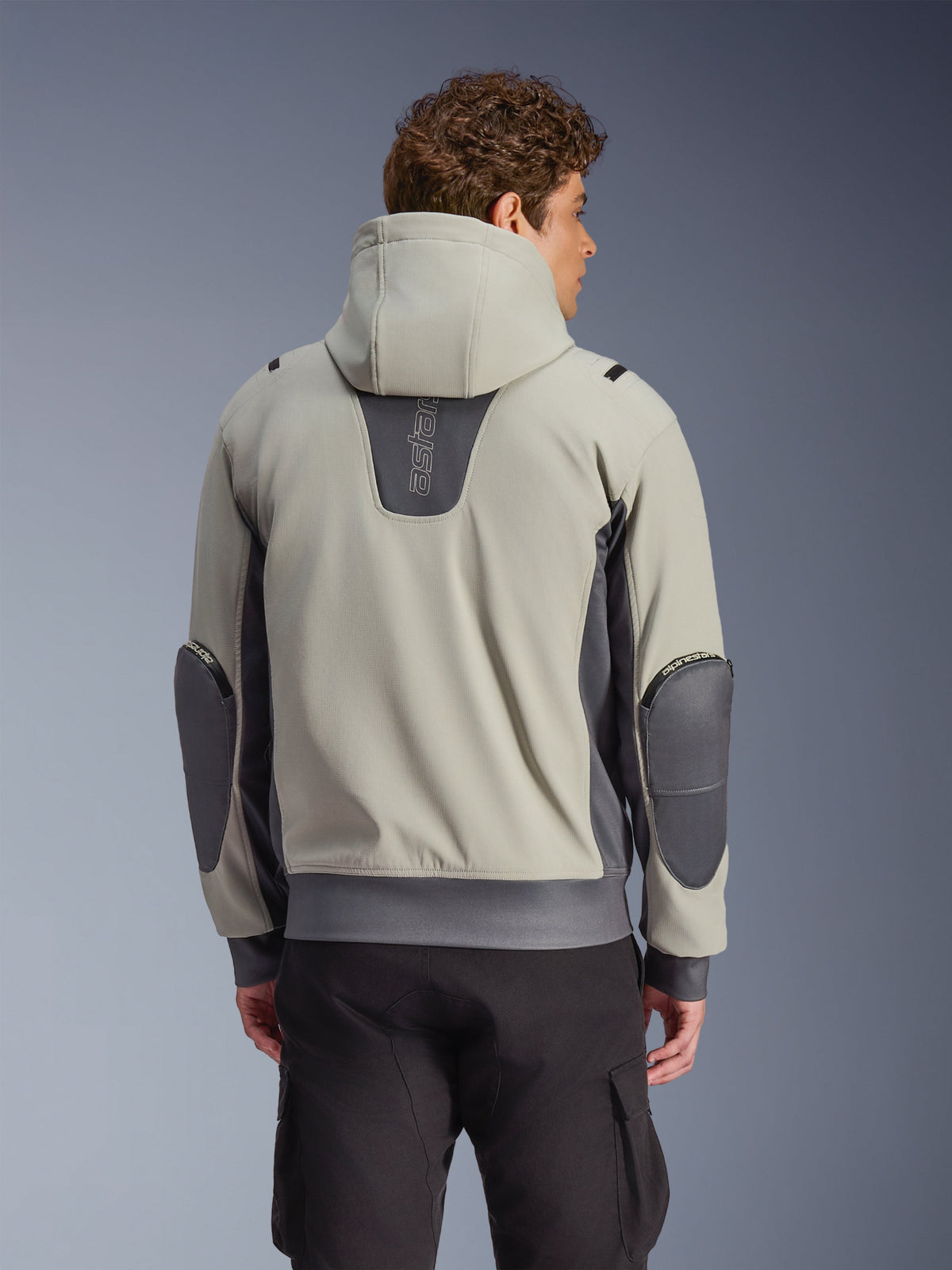 Alpinestars Moblast Waterproof Jas in Iron Gray en Dark Sage, achteraanzicht op model, stedelijke motorjas met capuchon voorzien van donkergrijze stretchpanelen en versterkte ellebogen, gecombineerd met een zwarte cargobroek