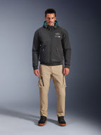 Alpinestars Moblast Waterproof Jacket, zwarte urban motorhoodie met groenblauwe binnenvoering in de capuchon, gedragen met een beige cargobroek en zwarte hoge boots, eigentijdse all-season rijkleding