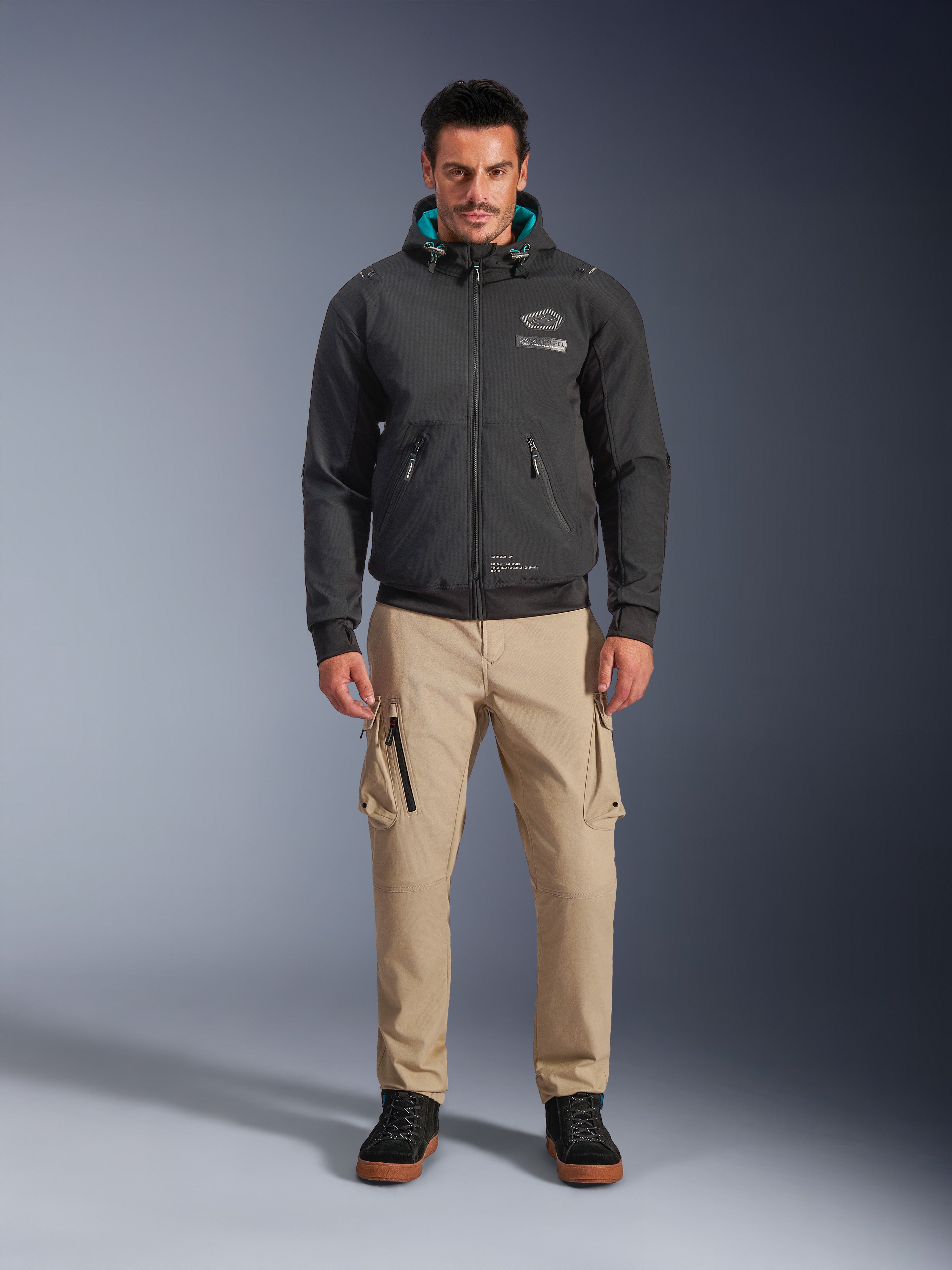 Alpinestars Moblast Waterproof Jacket, zwarte urban motorhoodie met groenblauwe binnenvoering in de capuchon, gedragen met een beige cargobroek en zwarte hoge boots, eigentijdse all-season rijkleding