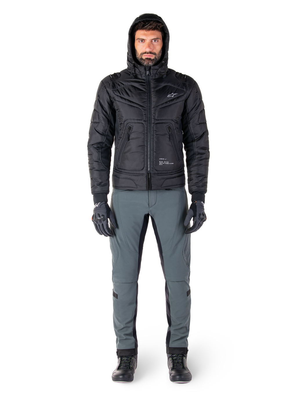 Alpinestars Mohobbs Waterproof Jas in zwart, mannelijk model draagt een gewatteerd bomberjack met capuchon, gecombineerd met een grijze motorbroek, zwarte handschoenen en zwarte laarzen, urban contemporary motoruitrusting