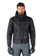 Alpinestars Mohobbs Waterproof Jacket, zwarte gewatteerde bomber motorjas, gewatteerde poly-stof met ritssluiting en ritszakken, klein wit logo op de borst, gedragen door een model met grijze broek en handschoenen, urban streetwear-ontwerp voor rijden in de stad