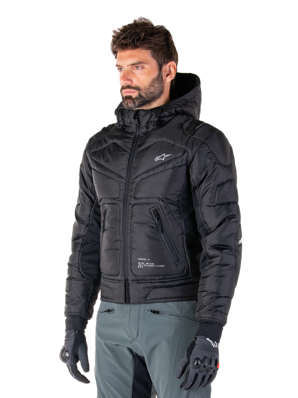 Alpinestars Mohobbs Waterproof Jacket, zwart, gewatteerd doorgestikt bombermodel met een capuchon, gedragen door een man met een grijze motorbroek en zwarte handschoenen, op urban streetwear geïnspireerde motorkleding met een klein wit logo op de borst.