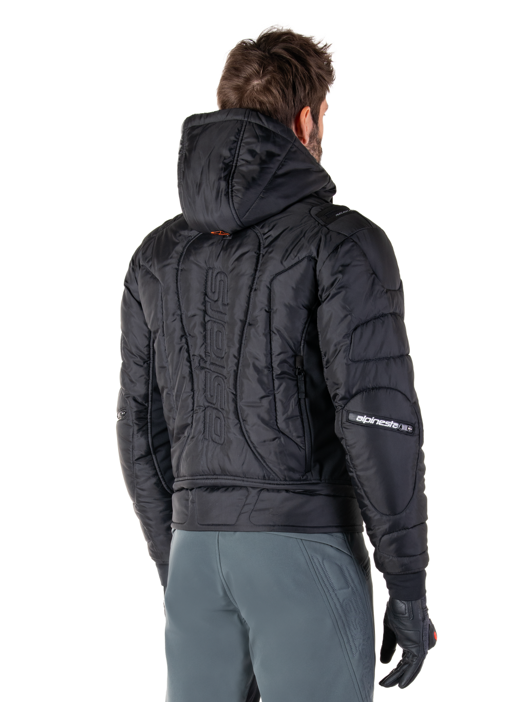 Alpinestars Mohobbs Waterproof jas, zwart, achteraanzicht met gewatteerde constructie, capuchon, verticaal astars-woordmerk op de rug en Alpinestars-branding op de mouwen, gedragen met grijze broek en zwarte handschoenen.