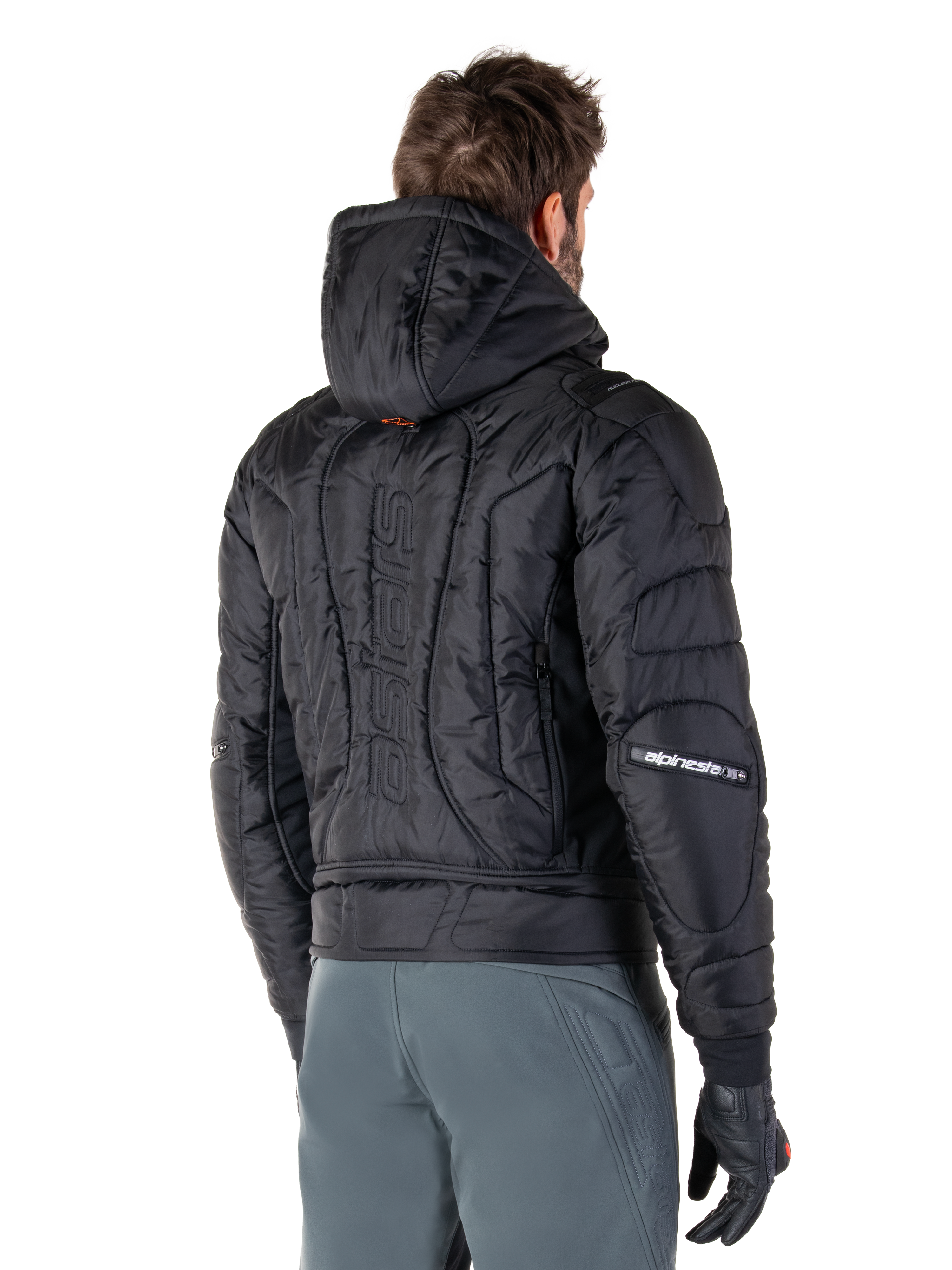 Alpinestars Mohobbs Waterproof jas, zwart, achteraanzicht met gewatteerde constructie, capuchon, verticaal astars-woordmerk op de rug en Alpinestars-branding op de mouwen, gedragen met grijze broek en zwarte handschoenen.
