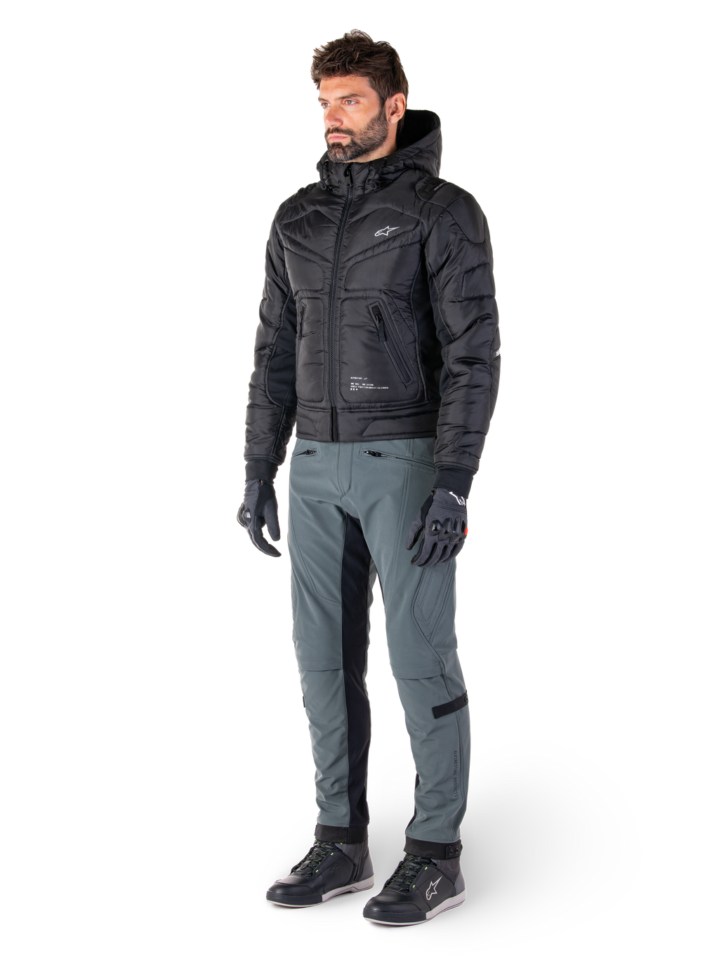 Alpinestars Mohobbs Waterproof Jacket, zwart, gewatteerde urban motorbomberjas met capuchon, gedragen door een model met grijze motorbroek en zwarte handschoenen, voorzien van een wit logo op de borst, eigentijdse op streetwear geïnspireerde beschermende kleding